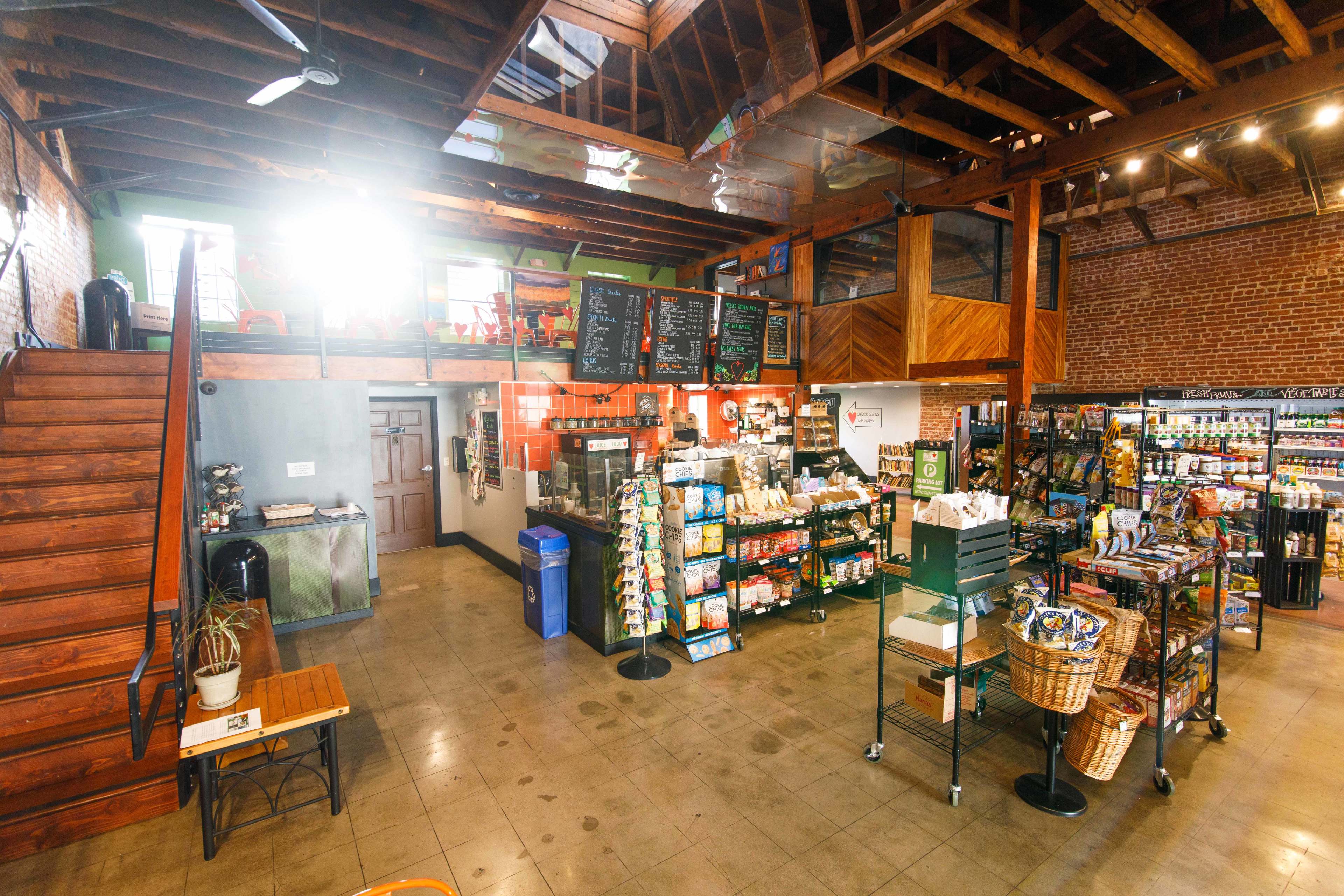 The interior of a market featuring a counter with various goods for sale, exposed brick walls, and a staircase leading to an upper level.