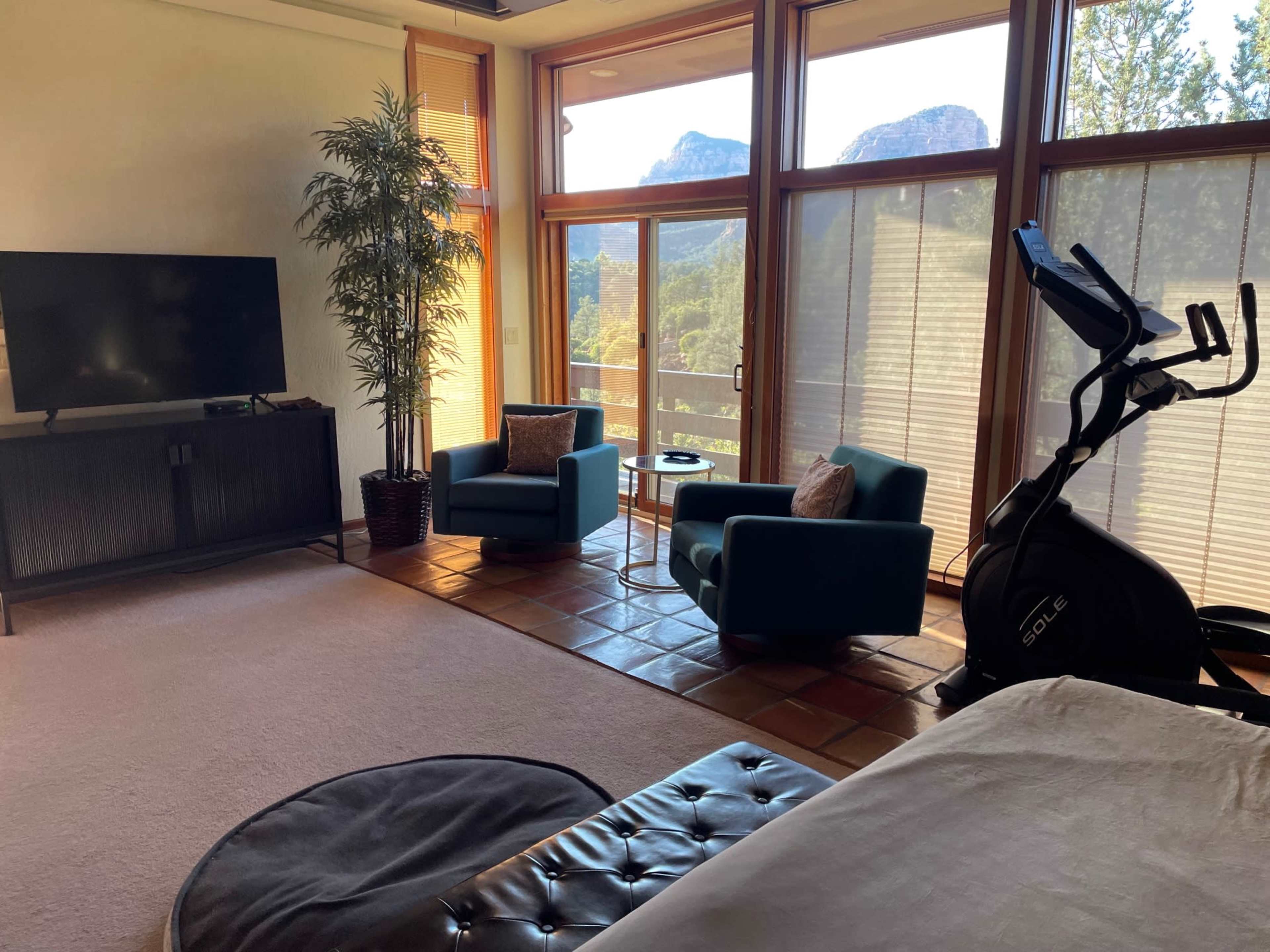 A living area with two blue chairs, a small table, a television unit, an elliptical machine, and large windows revealing a mountainous view.