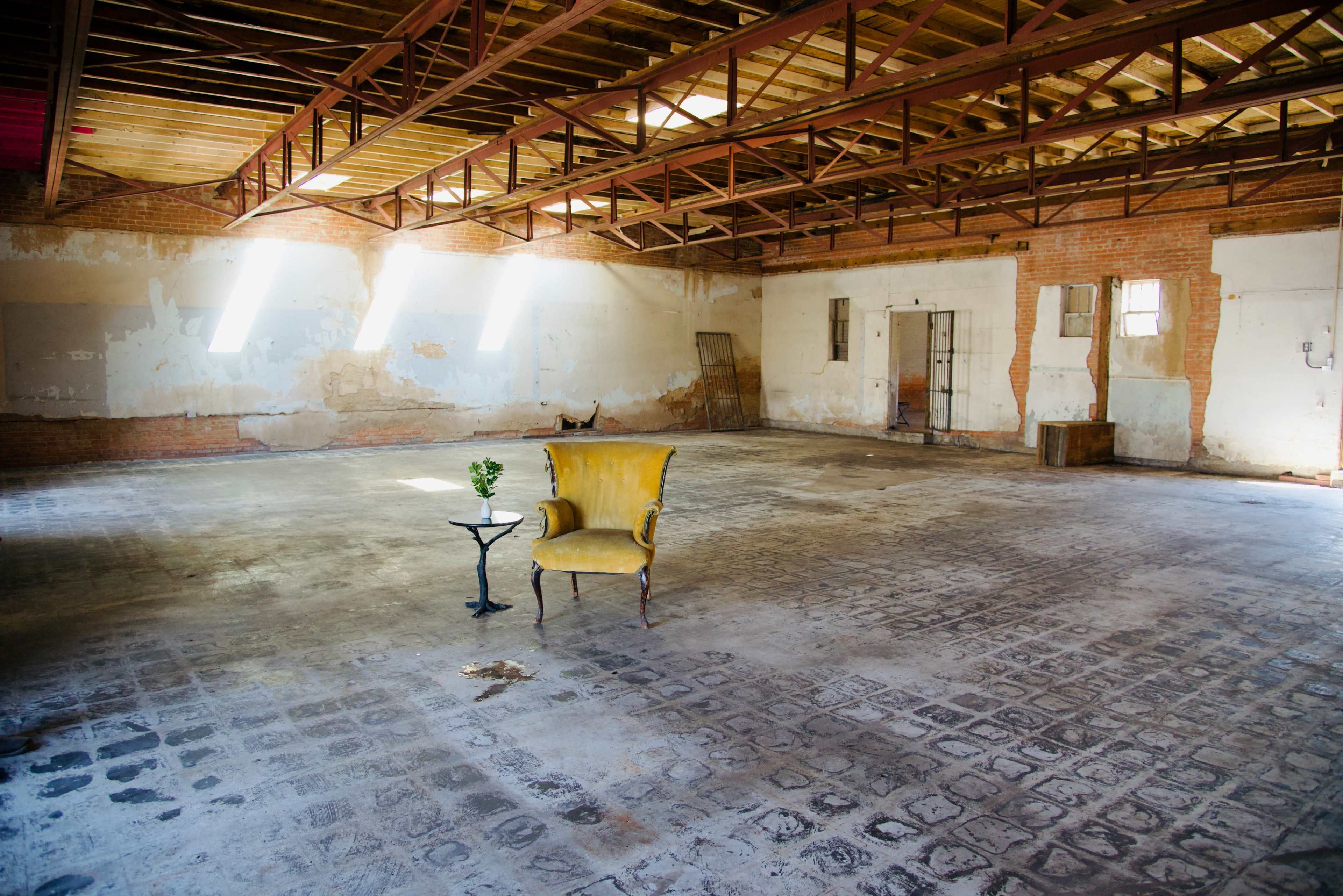 An empty industrial space features a single yellow chair and a small table with a plant in the center.