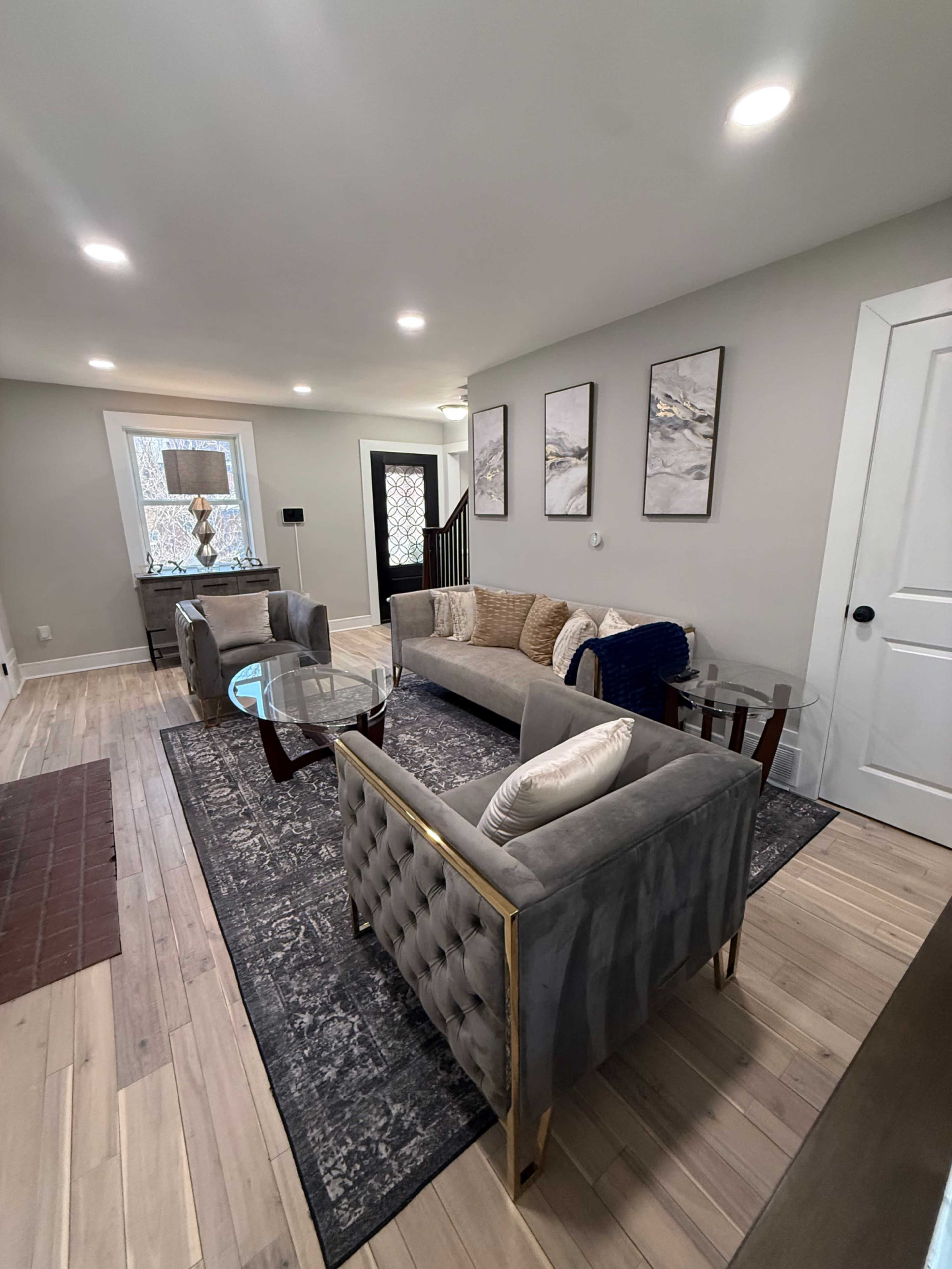 The image shows a modern living room with a gray sectional sofa, two glass-top tables, and framed artwork on the walls.