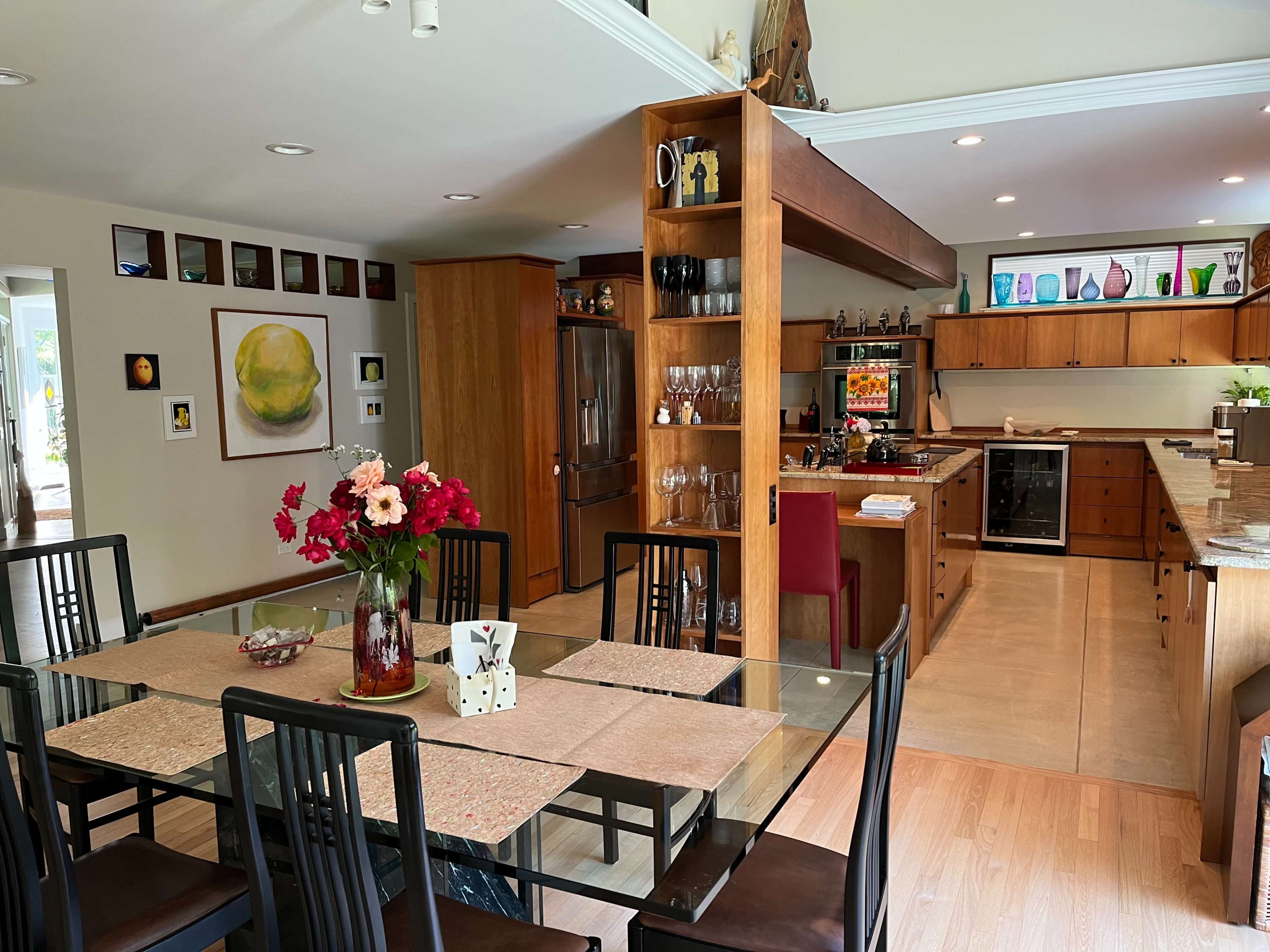 A modern kitchen and dining area featuring a wooden dining table set with flowers, surrounded by black chairs, and an open kitchen with wooden cabinetry and various glassware displayed.