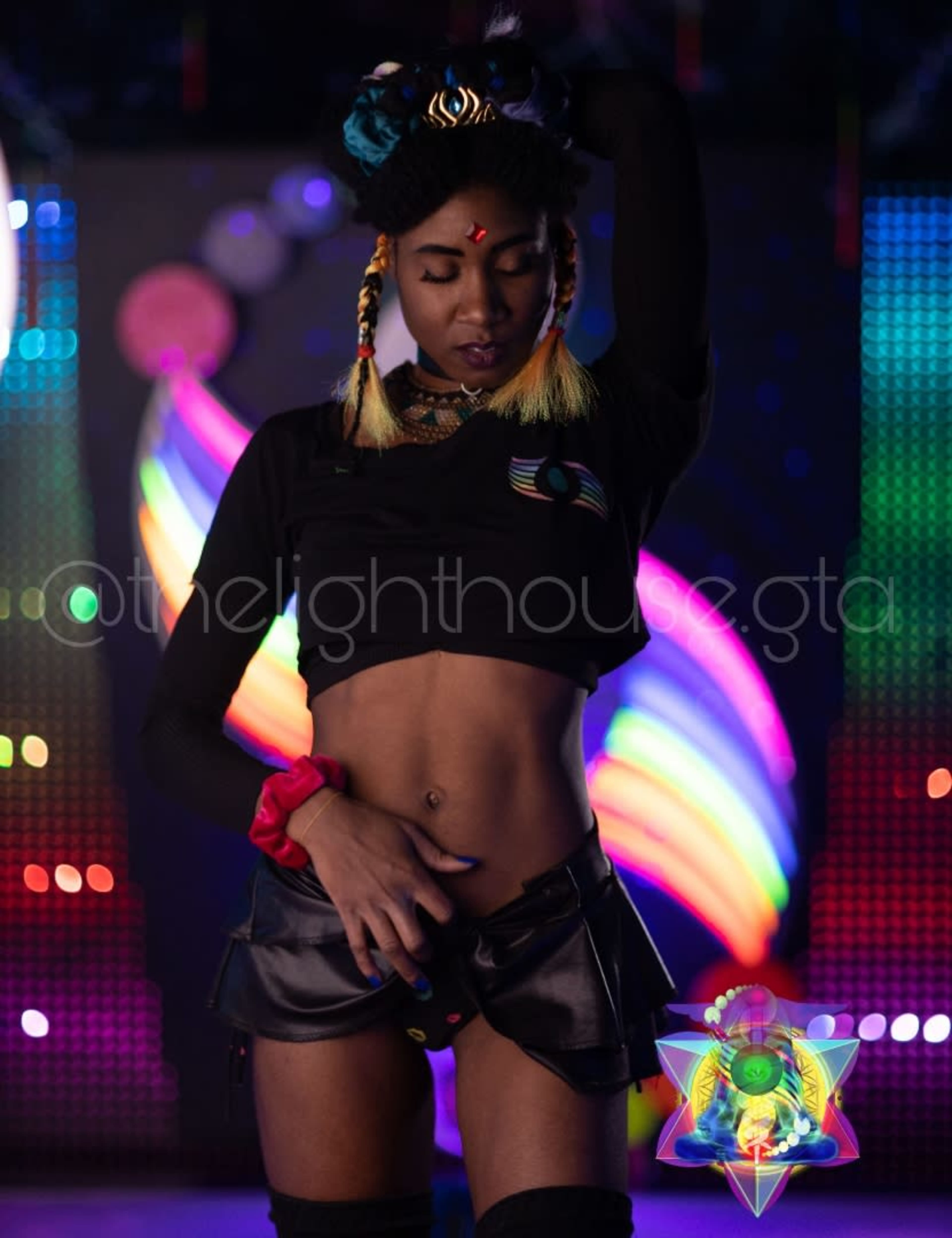 A performer in a black crop top and shiny shorts poses on stage with colorful lights in the background.