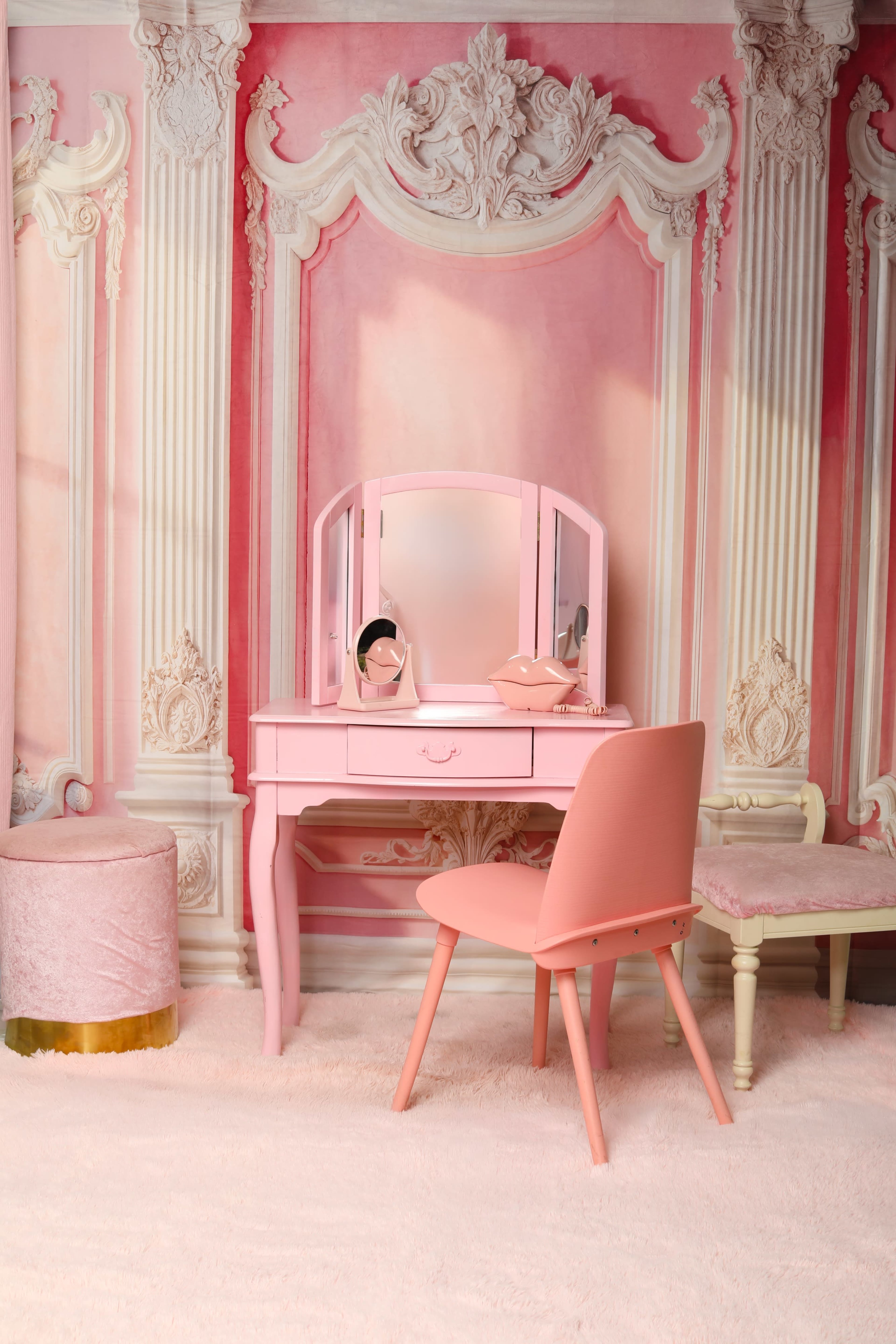 The image features a pink vanity table with a mirror and a matching chair, set against a pink and white decorative wall with ornate moldings.
