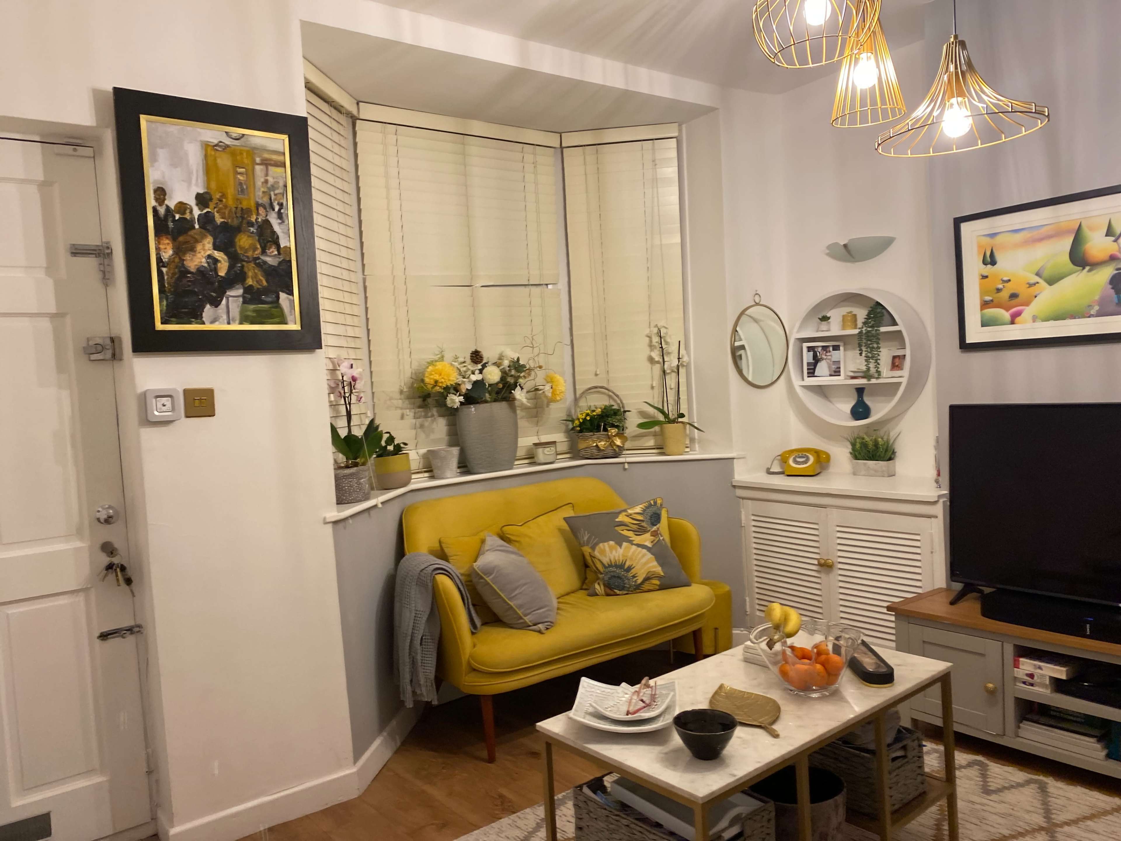 A cozy living room features a yellow sofa, round coffee table with fruit, and a wall adorned with art and decorative items.