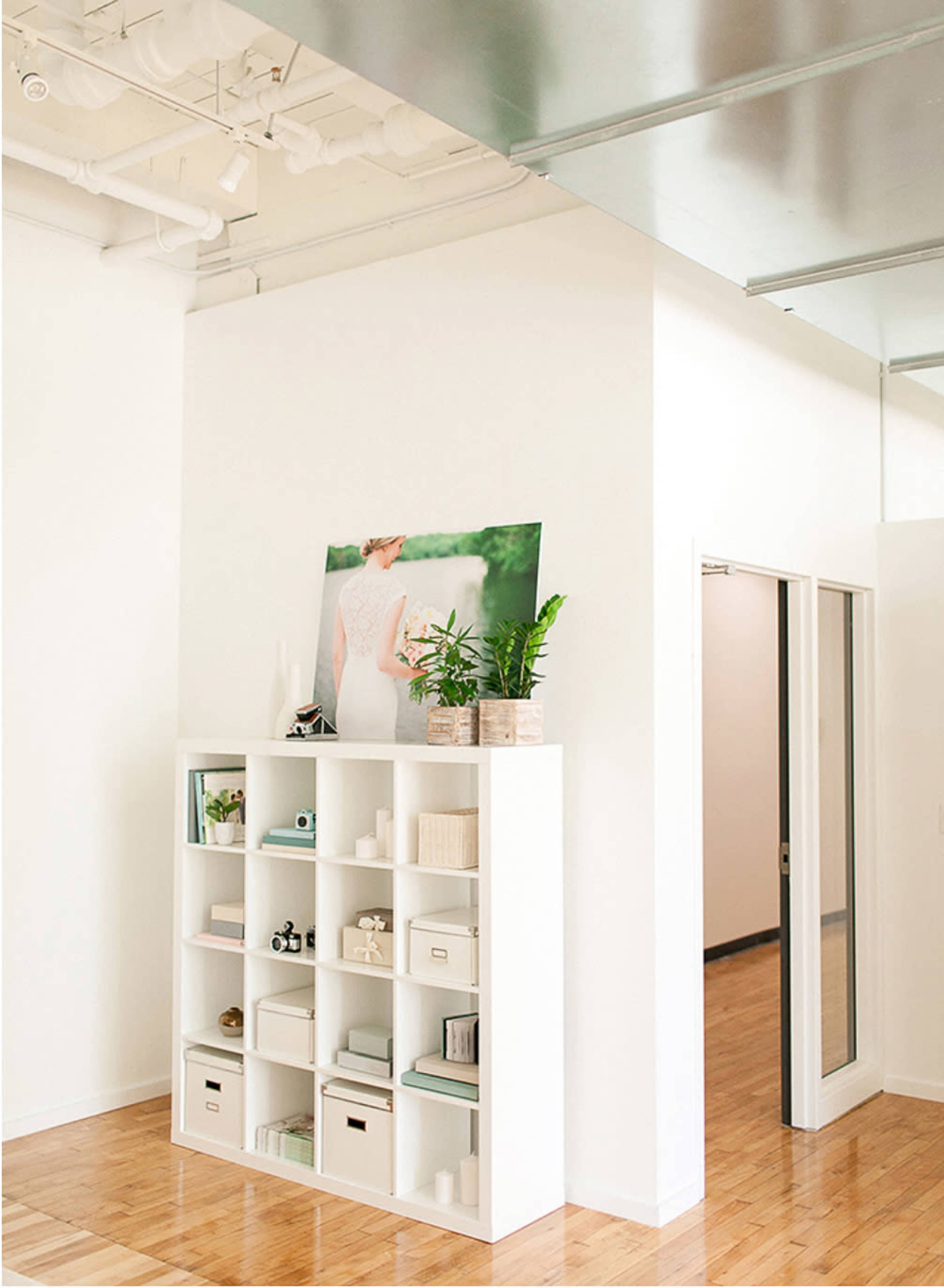 The image shows a bright, modern room with a white bookshelf filled with various containers and decor, a large photo on the wall, and a doorway leading to another space.