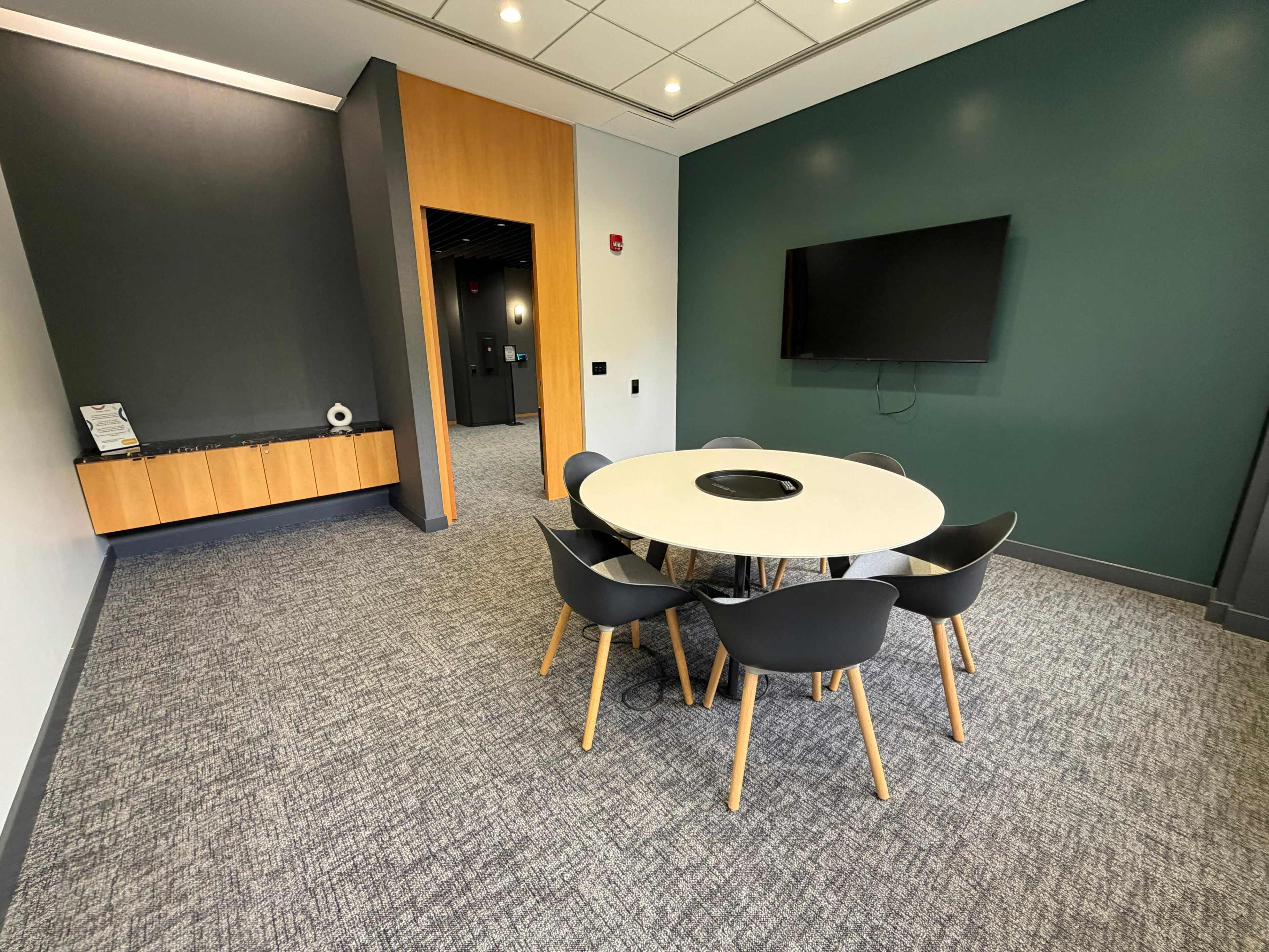 The image shows a modern conference room with a round table surrounded by gray chairs, a television mounted on the wall, and a side cabinet against a dark green accent wall.