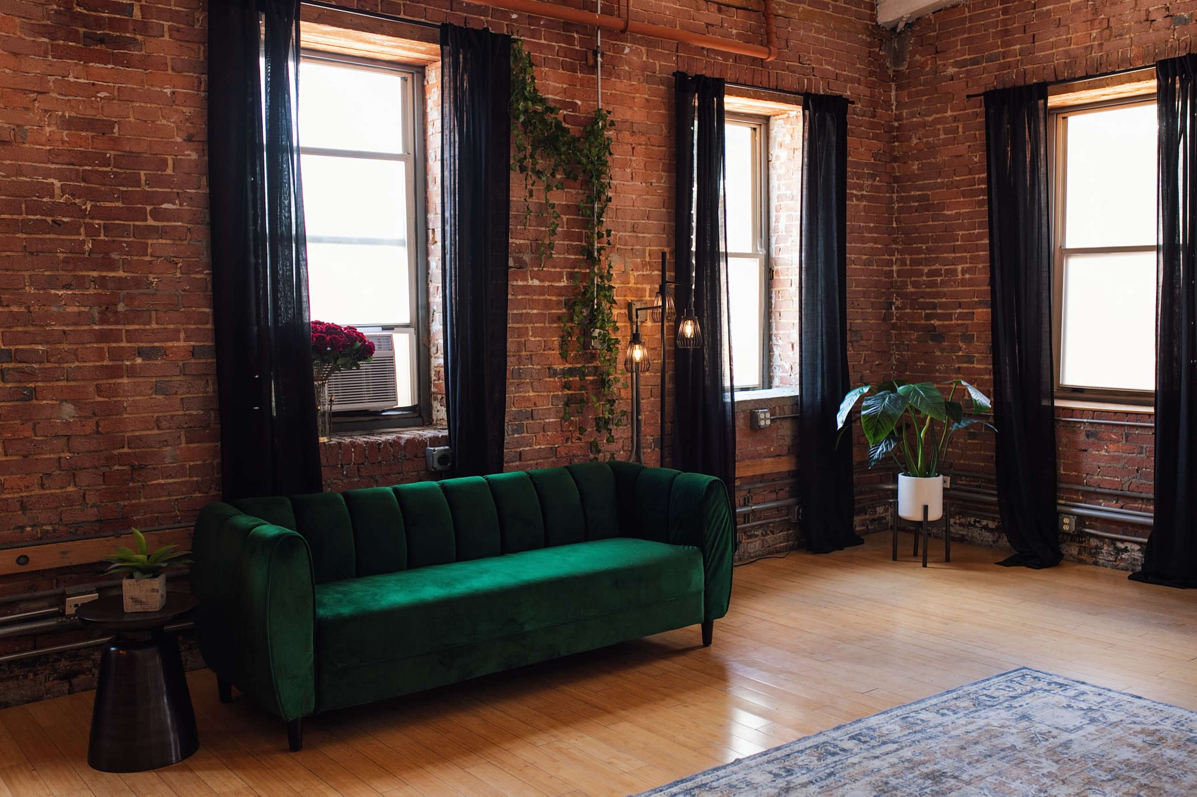 The image shows a green velvet sofa facing two windows in a room with exposed brick walls, wooden floors, and decorative plants.