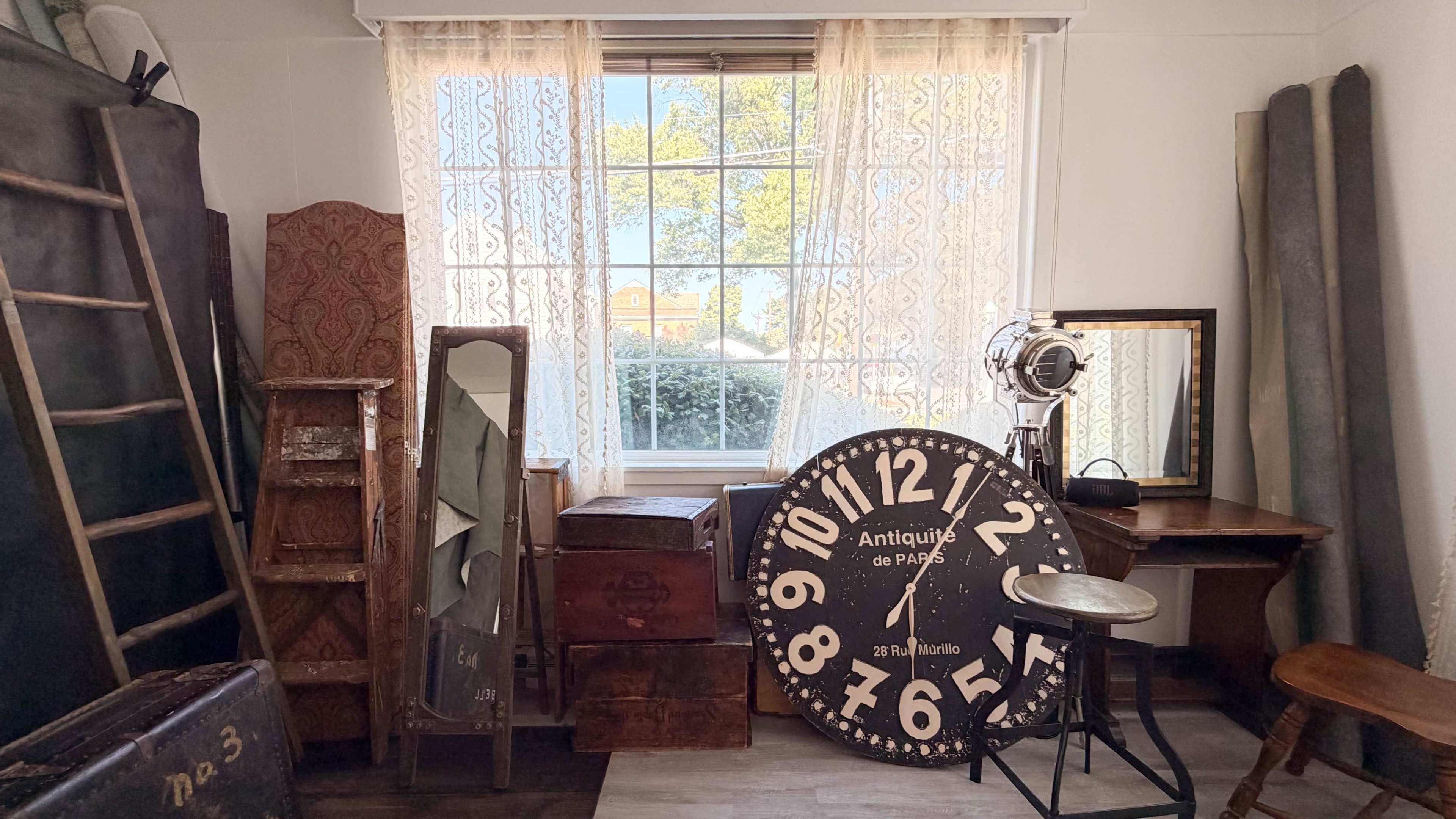 The scene depicts an interior room filled with various antique furniture pieces, including a large wall clock, mirrors, and a wooden desk, illuminated by natural light filtering through a window with lace curtains.