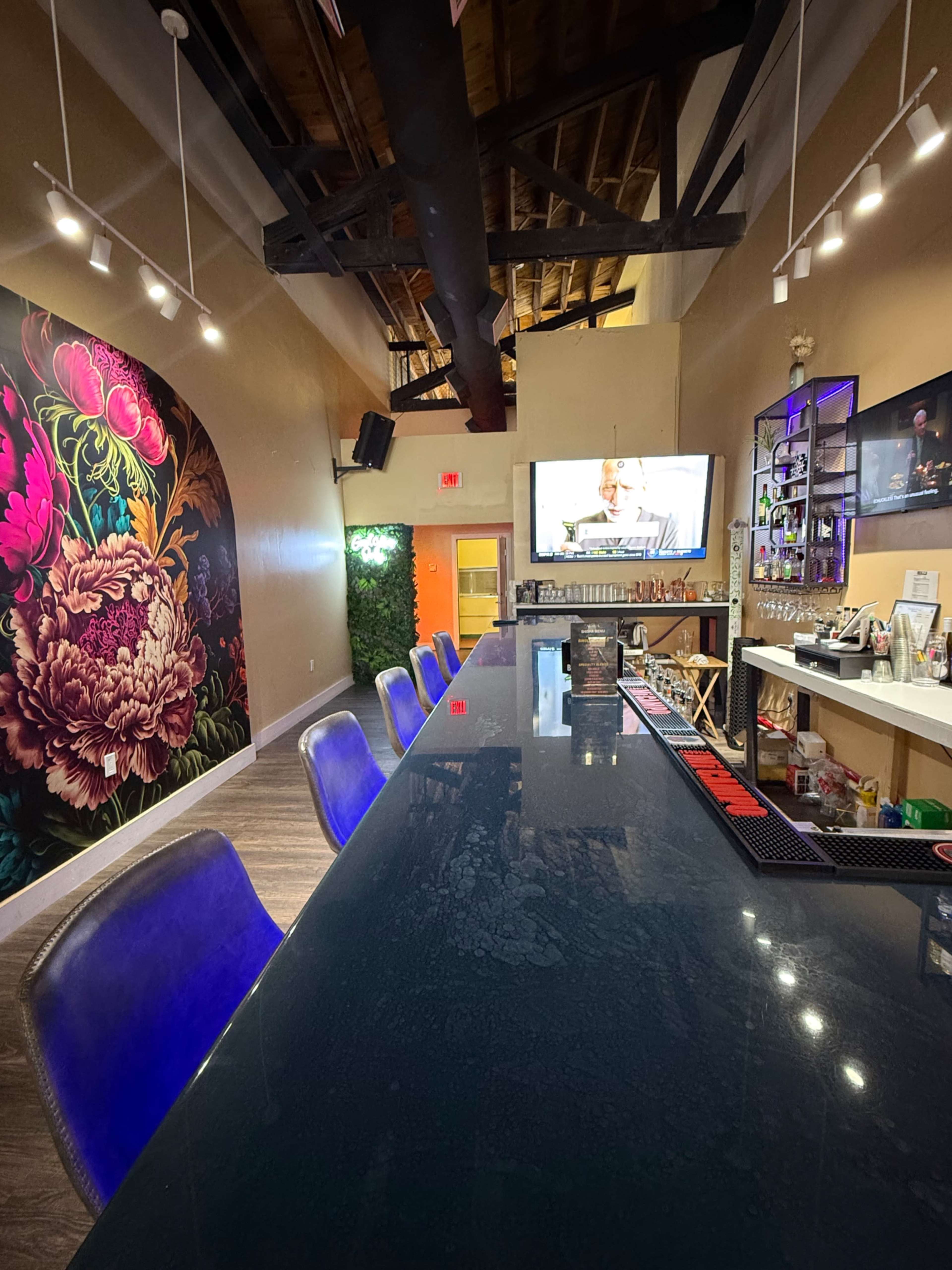 The image shows a modern bar area with a glossy counter, colorful wall art, a television, and a shelf of drinks behind the counter.