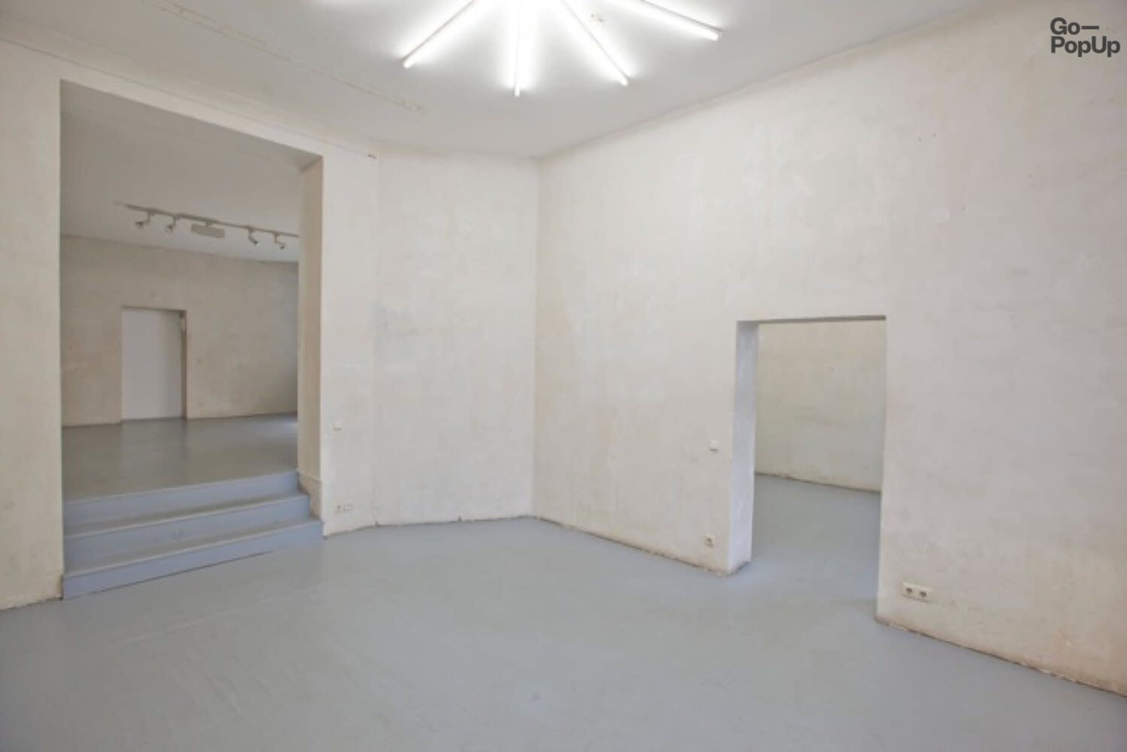 An empty, well-lit room with light gray flooring and plain, white walls, featuring doorways leading to adjacent spaces.