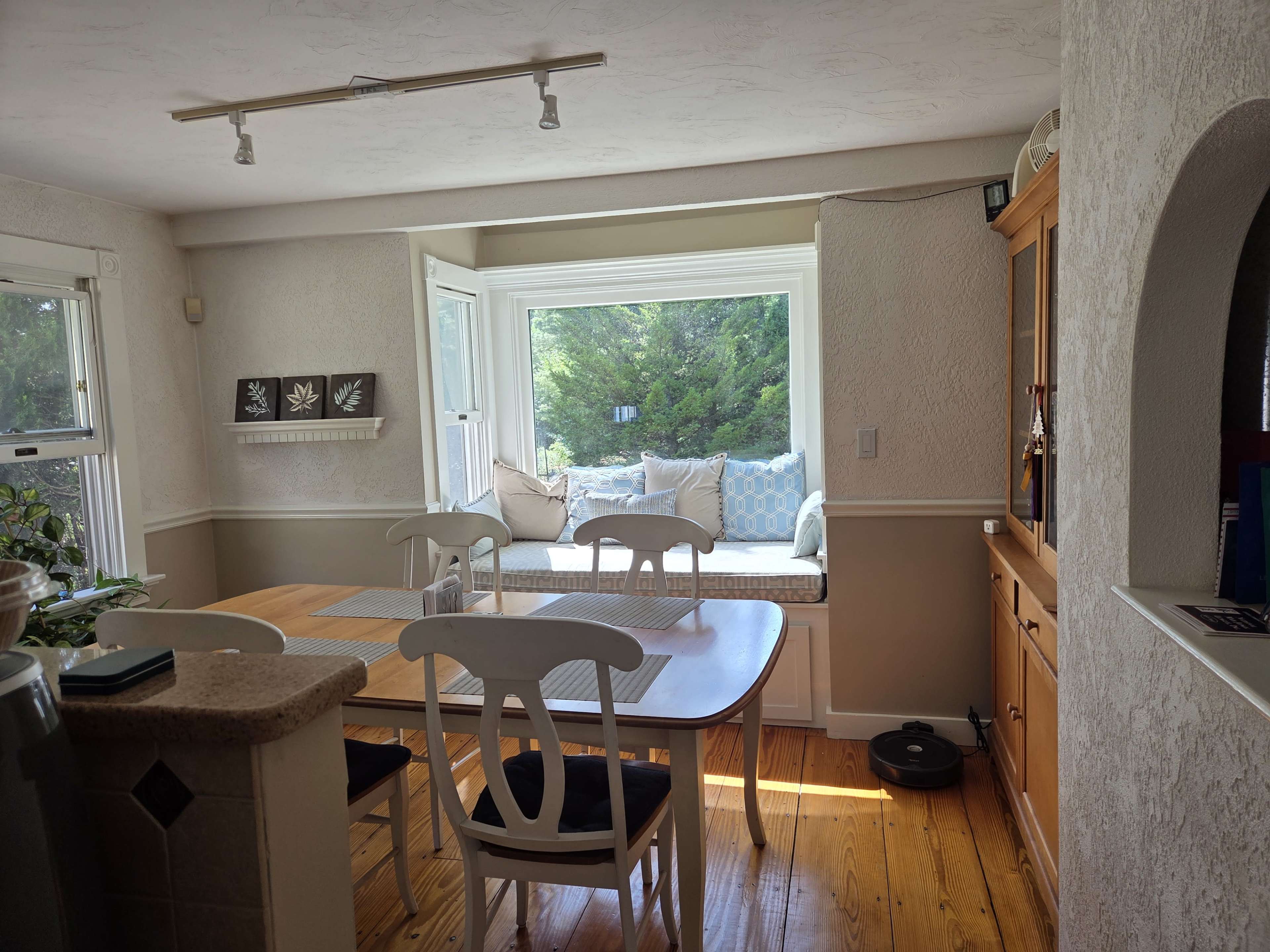 A bright kitchen features a dining table and chairs, with a window nook displaying cushions and greenery outside.