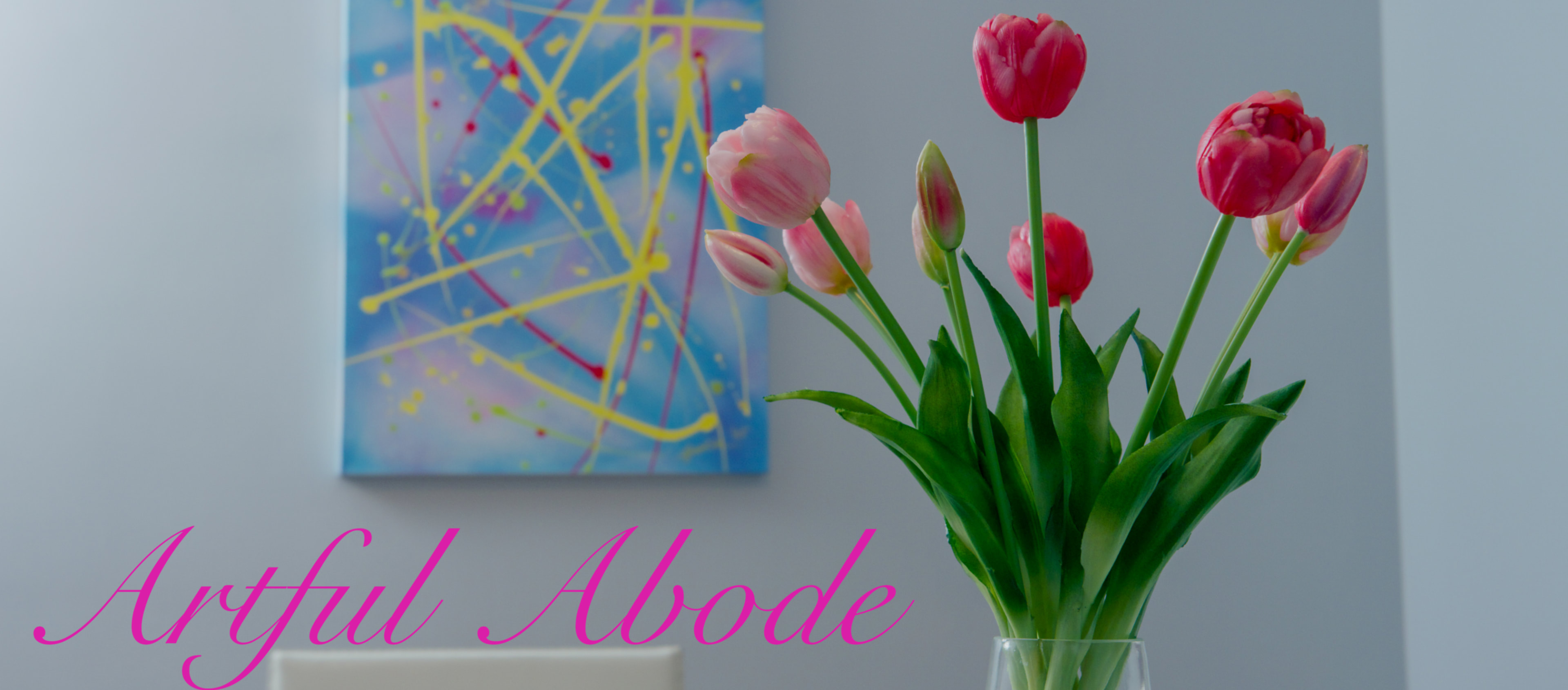 A vase filled with pink tulips stands in front of an abstract painting featuring bright colors and dynamic lines.