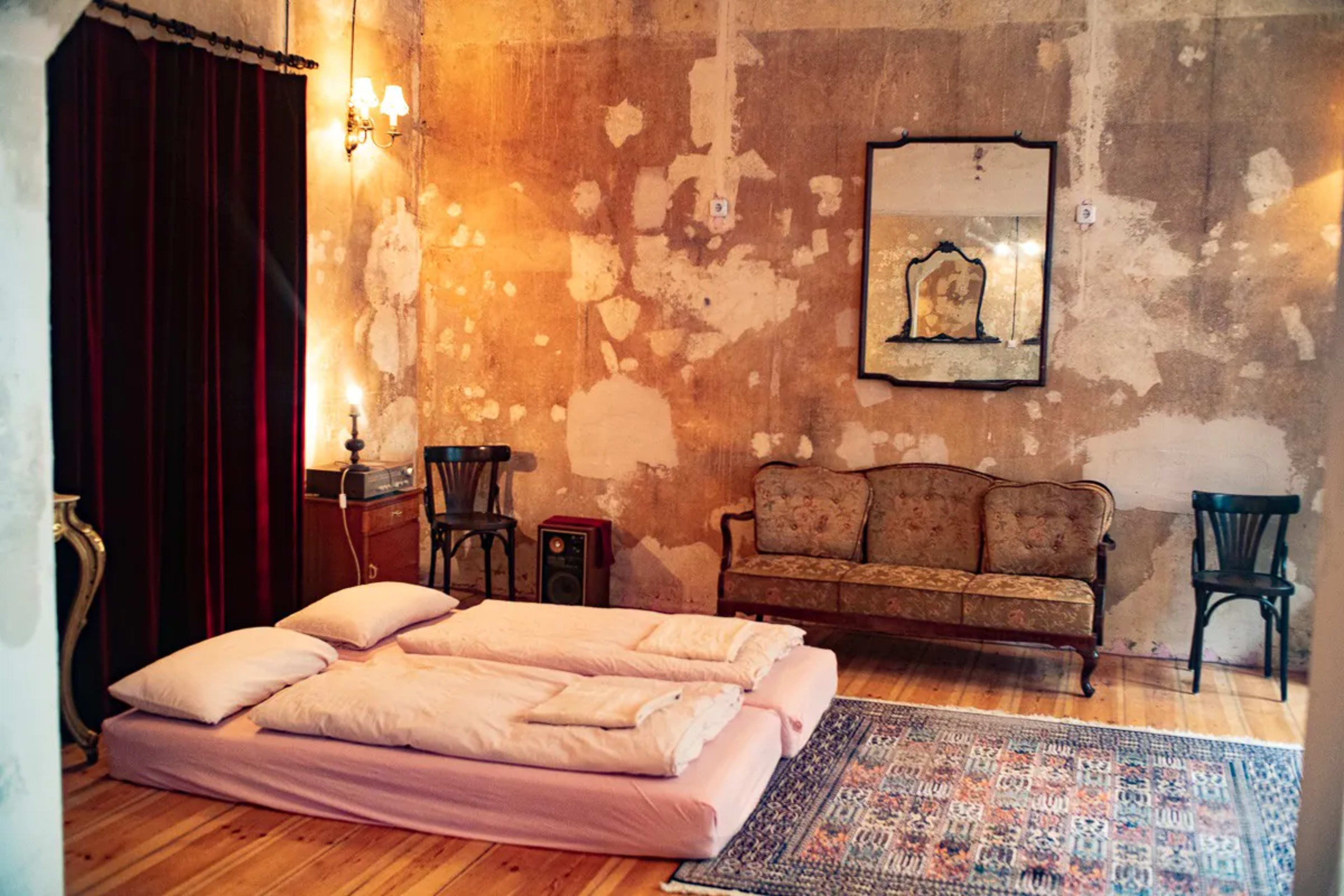 A sparsely furnished room with a simple bedding arrangement on the floor, a vintage sofa, a side table, and a patterned rug on a wooden floor, all set against a textured, peeling wall.