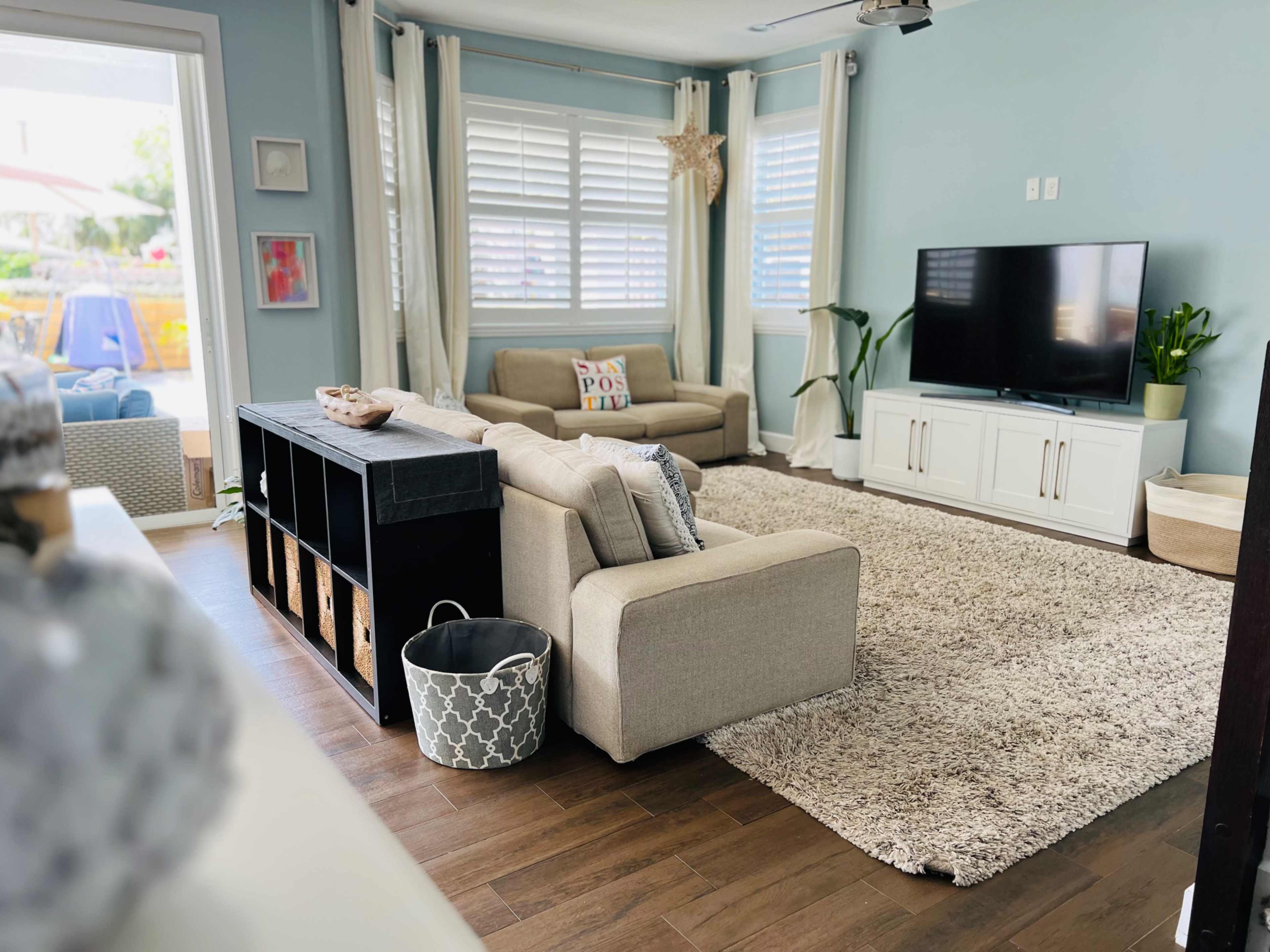 The image shows a bright living room with light blue walls, a large rug, a sectional sofa, a television stand, and potted plants.