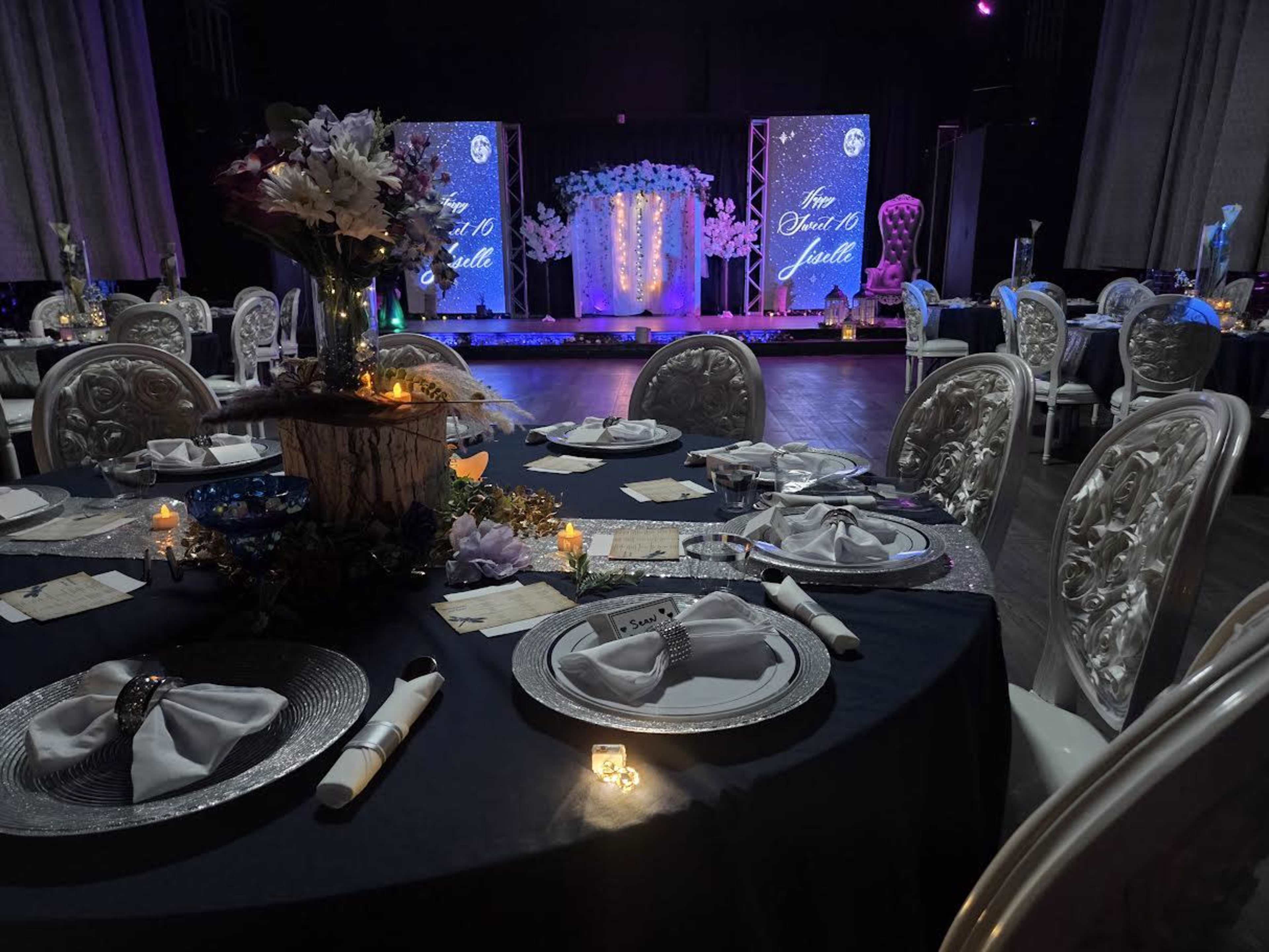 A banquet hall is set with elegantly arranged tables covered in dark tablecloths, featuring decorative centerpieces, plates, and lit candles, with a stage adorned with floral decorations in the background.
