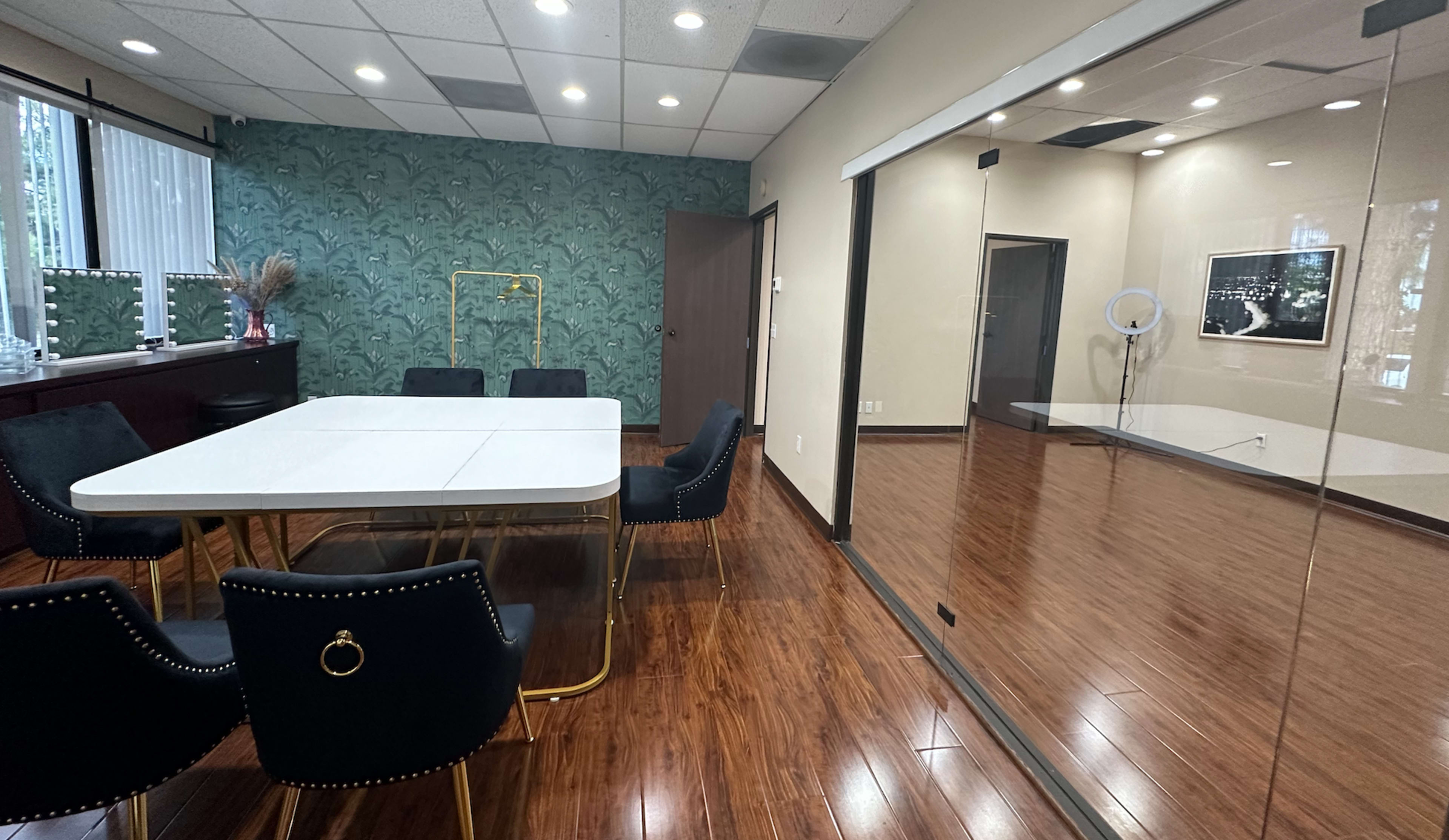 A meeting room with a white table surrounded by black chairs, featuring a green patterned wallpaper and a mirrored wall reflecting the space.
