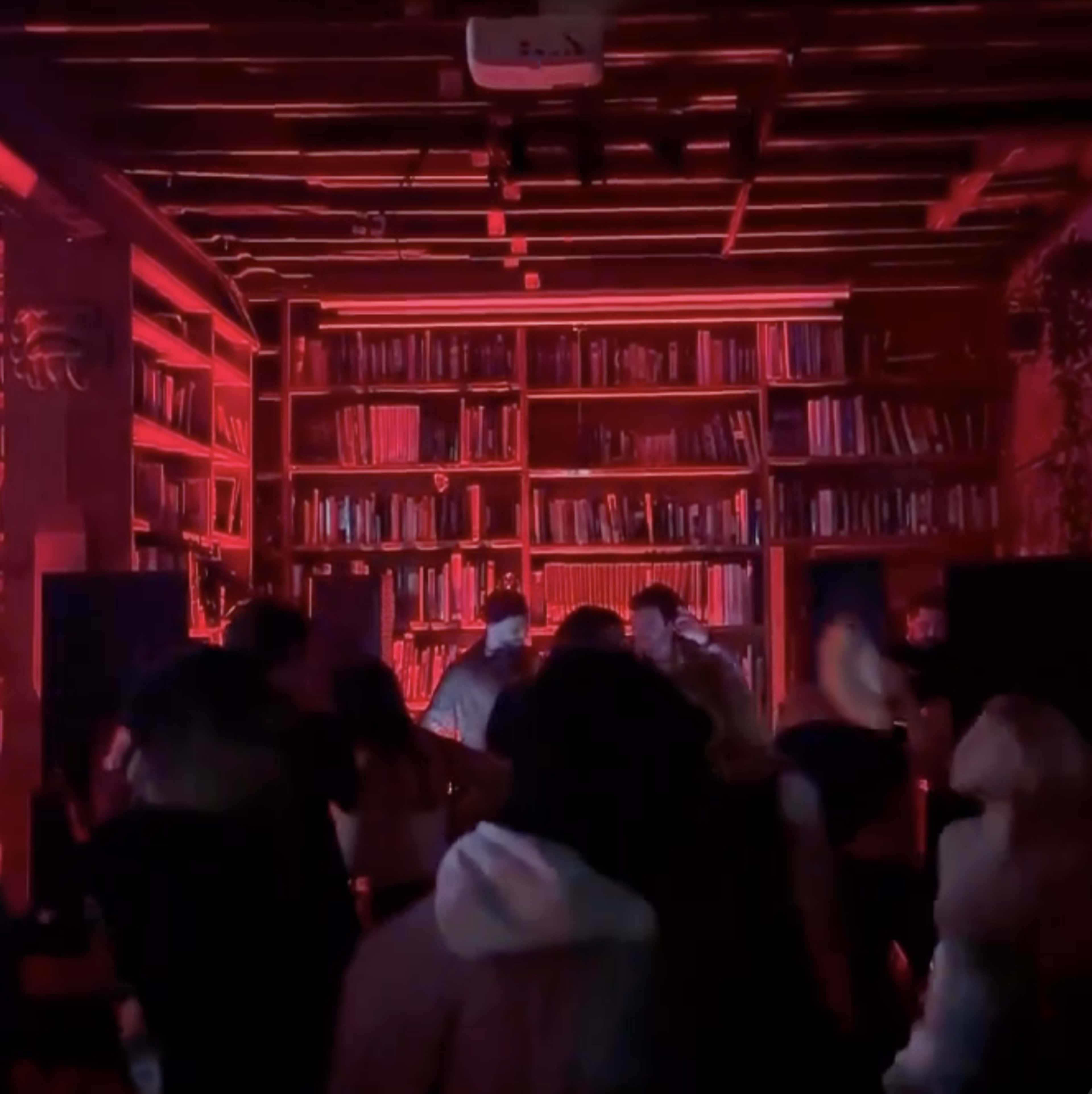 A group of people dance in front of a DJ setup surrounded by shelves of books illuminated by red lighting.