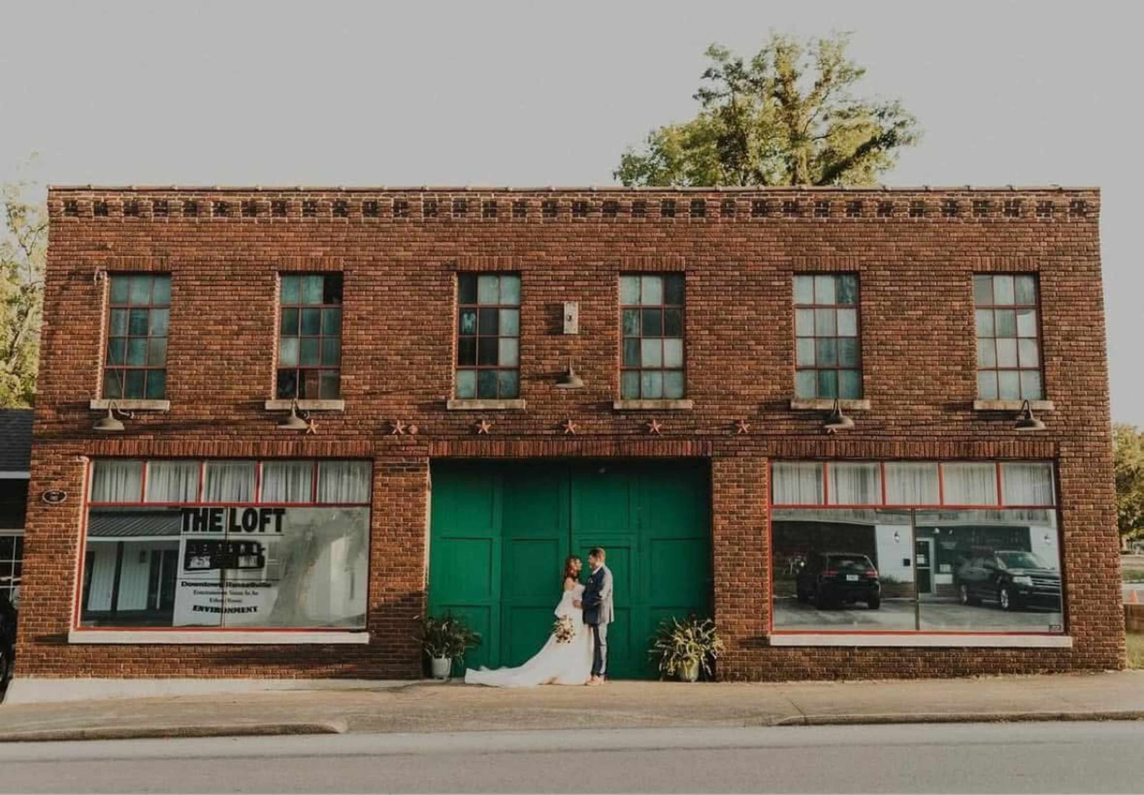 A couple in wedding attire stands in front of a brick building with green doors and large windows.