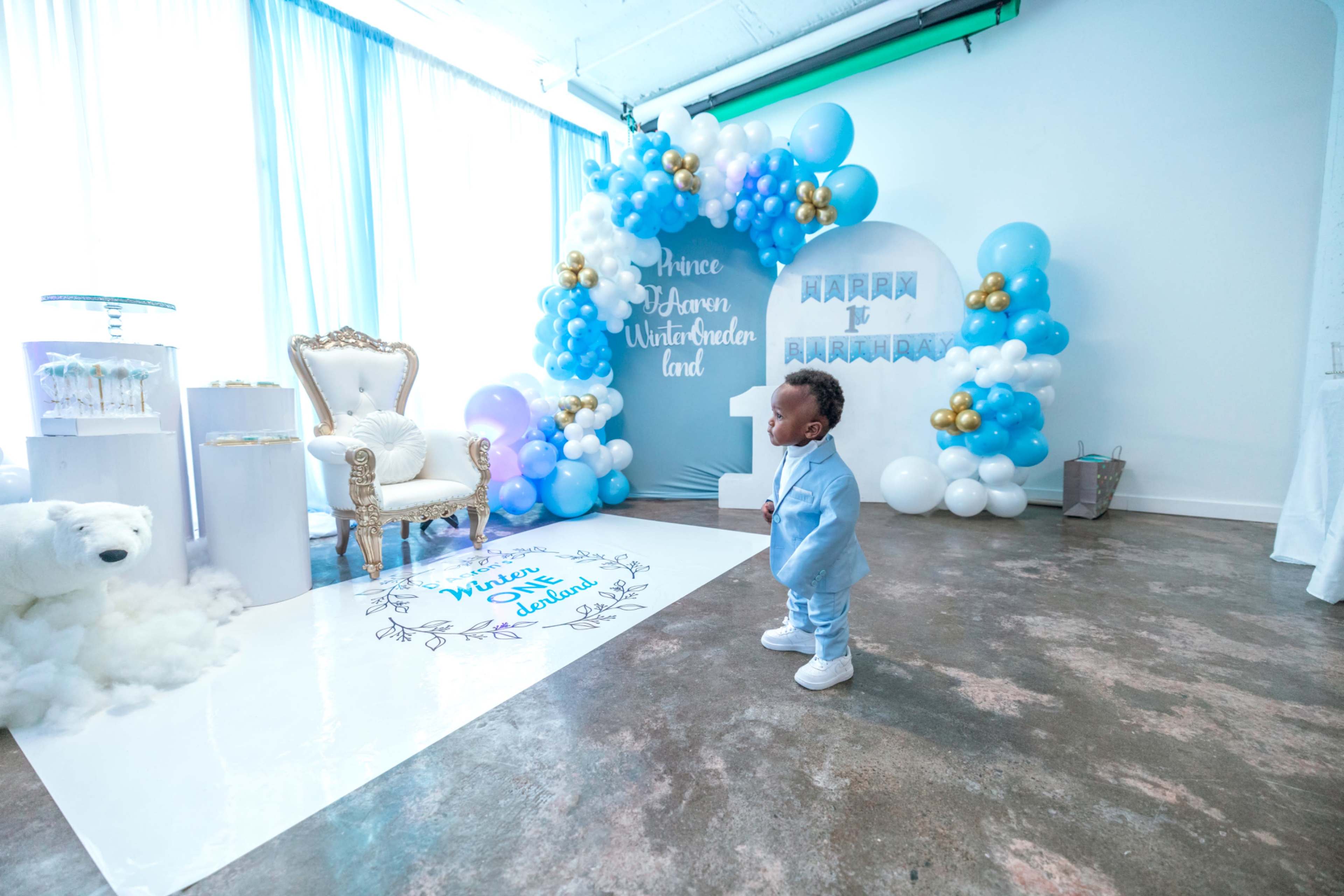 A child in a light blue outfit stands on a white carpet in a decorated room featuring balloons and a backdrop for a winter-themed celebration.