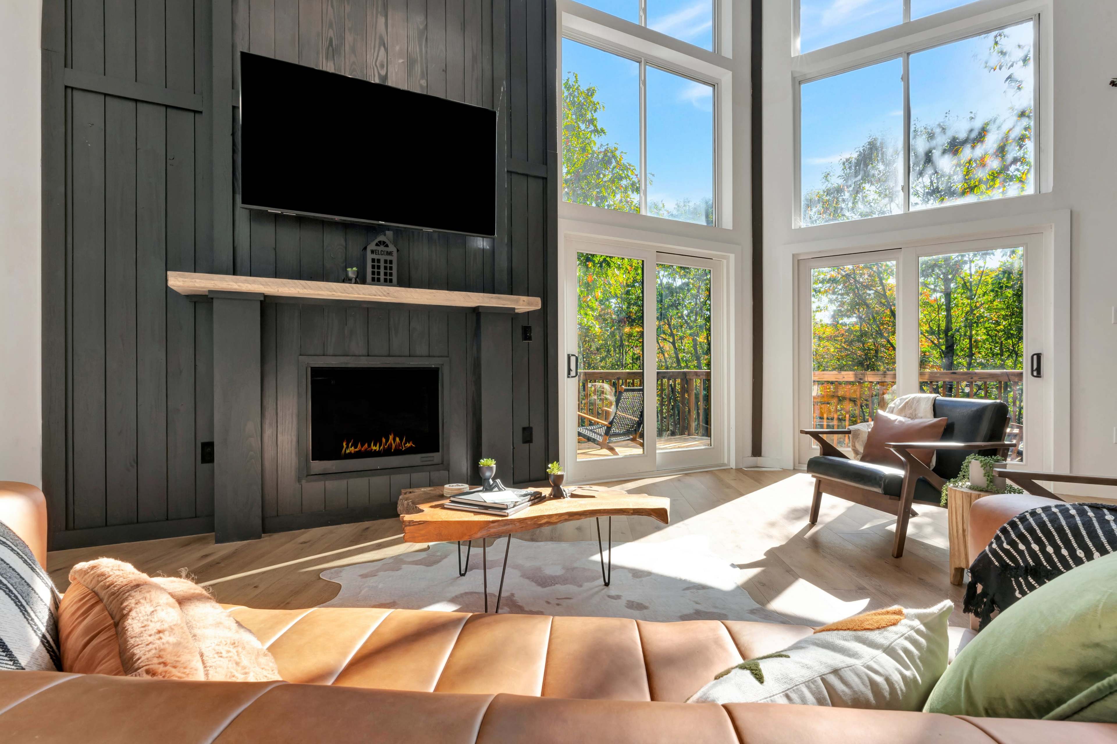 The image shows a modern living room with a large television mounted above a fireplace, large windows illuminating the space, and stylish furniture including a brown leather sofa and a wooden coffee table.