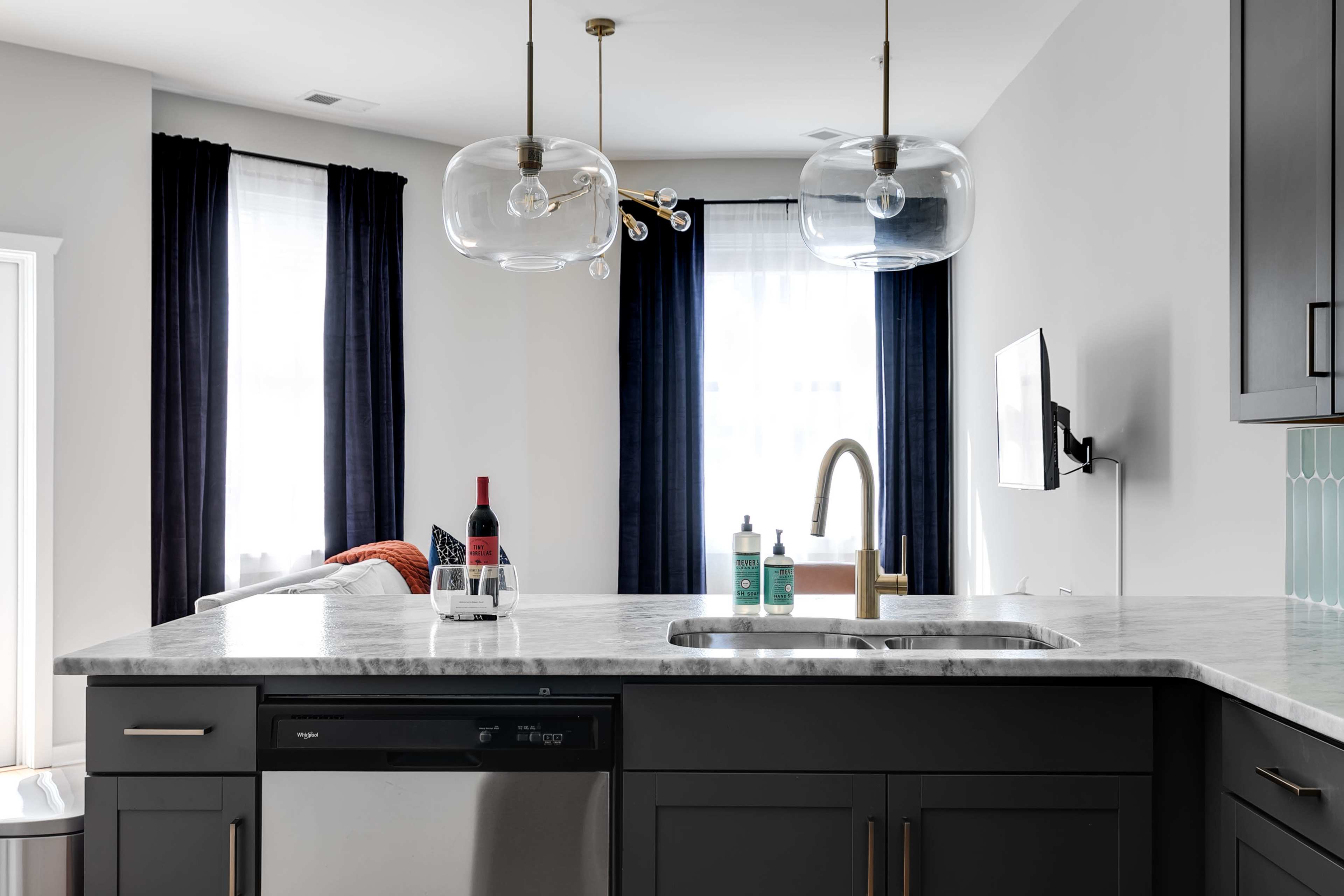 A modern kitchen features a marble countertop, dark cabinetry, and pendant lights above the sink, with bottles and a wine glass on the surface.
