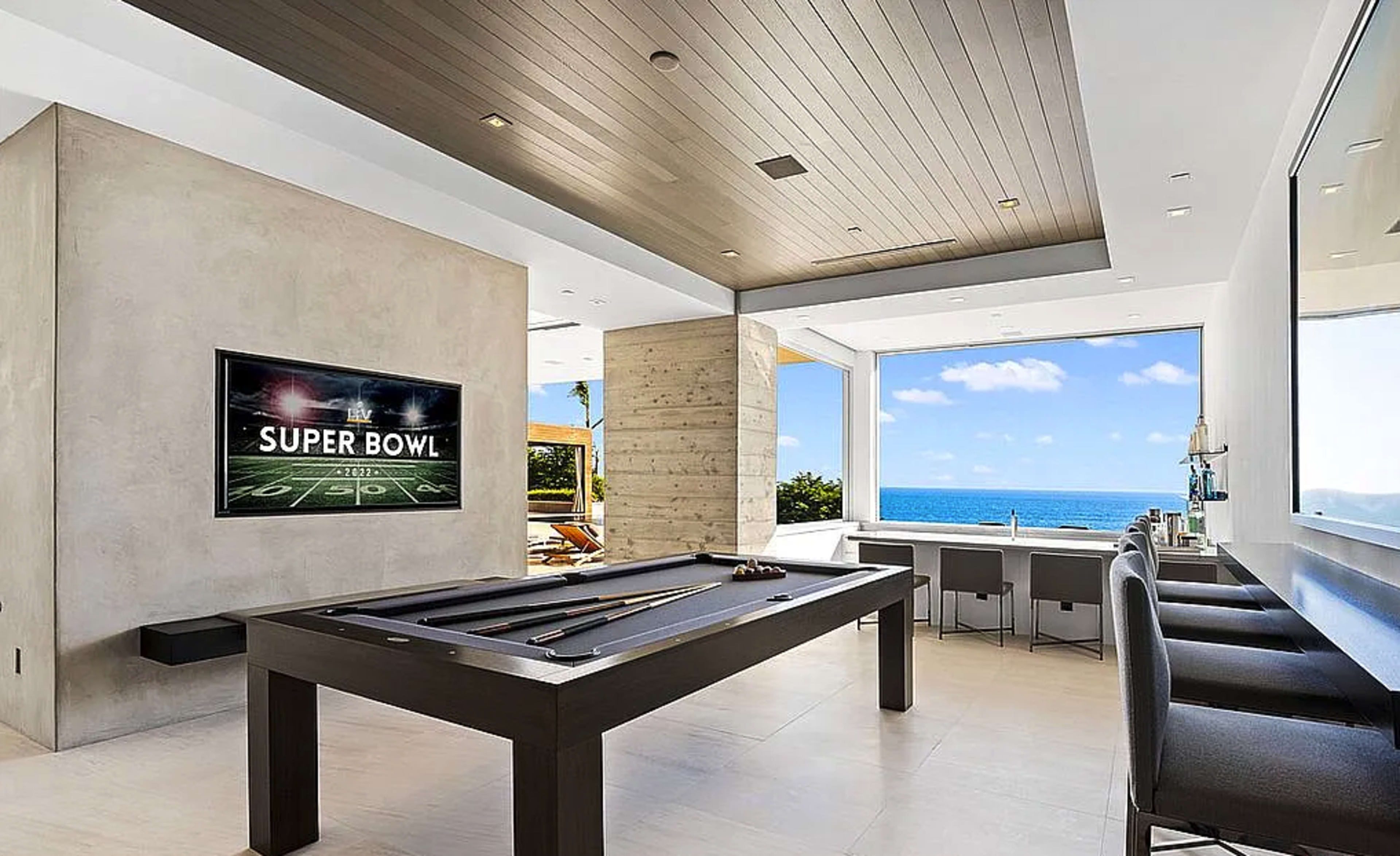 The image shows a modern living space featuring a billiard table, a large wall-mounted television displaying "SUPER BOWL," and a panoramic ocean view.