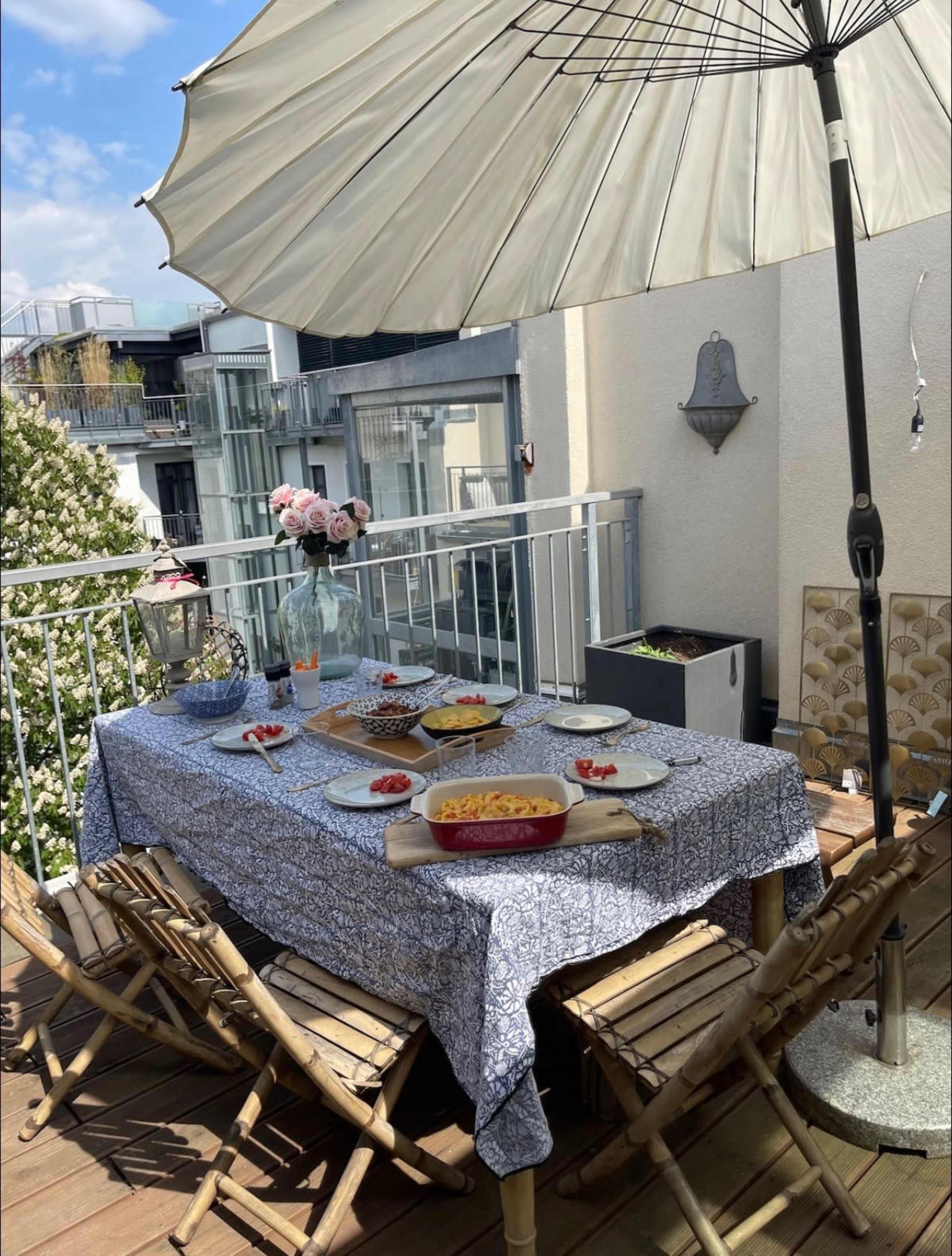 A table set for a meal is placed on a balcony, featuring a tablecloth, various dishes, and an umbrella for shade.