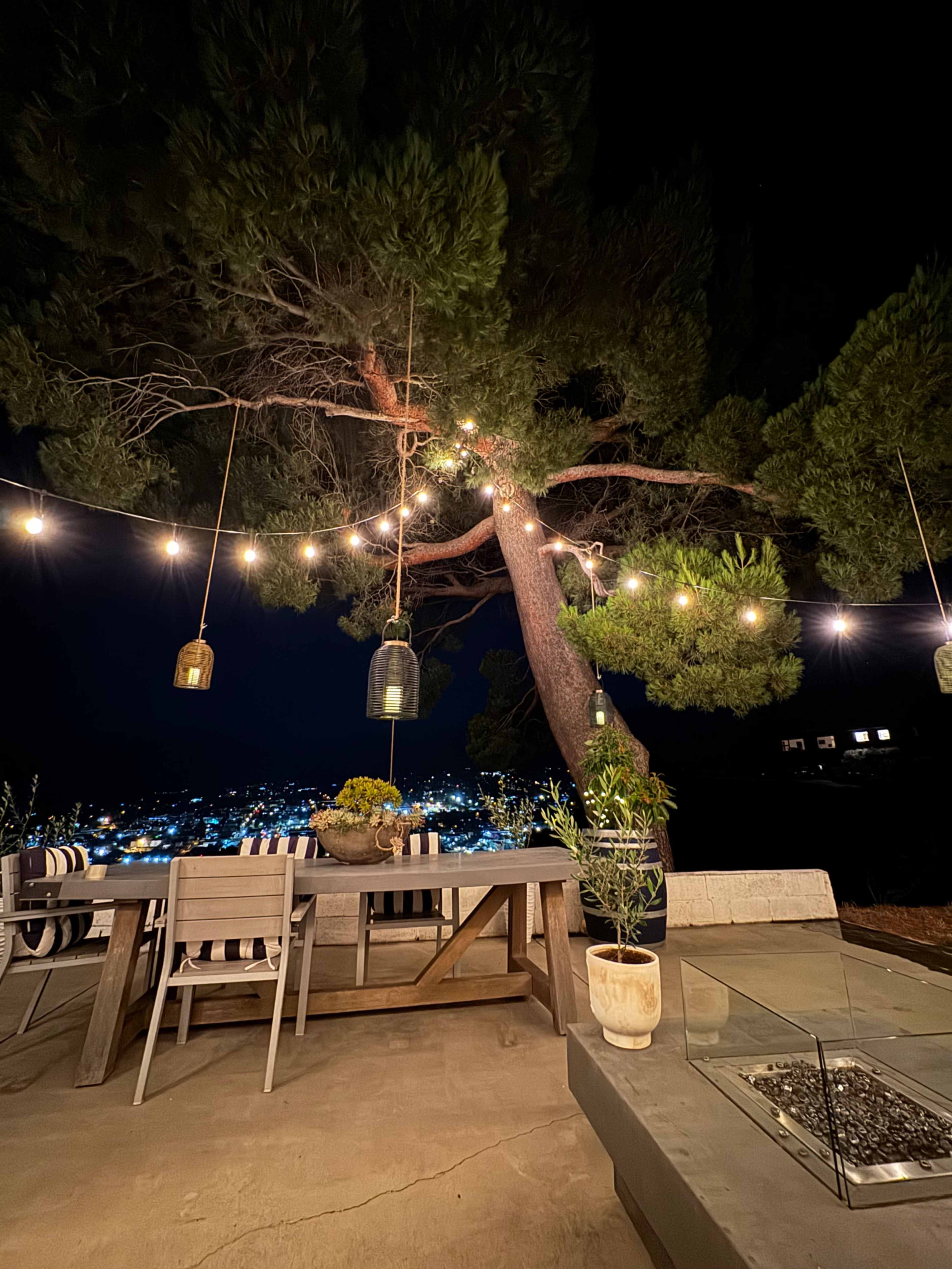 A patio table with chairs is set under a large tree adorned with hanging lights, overlooking a city illuminated at night.