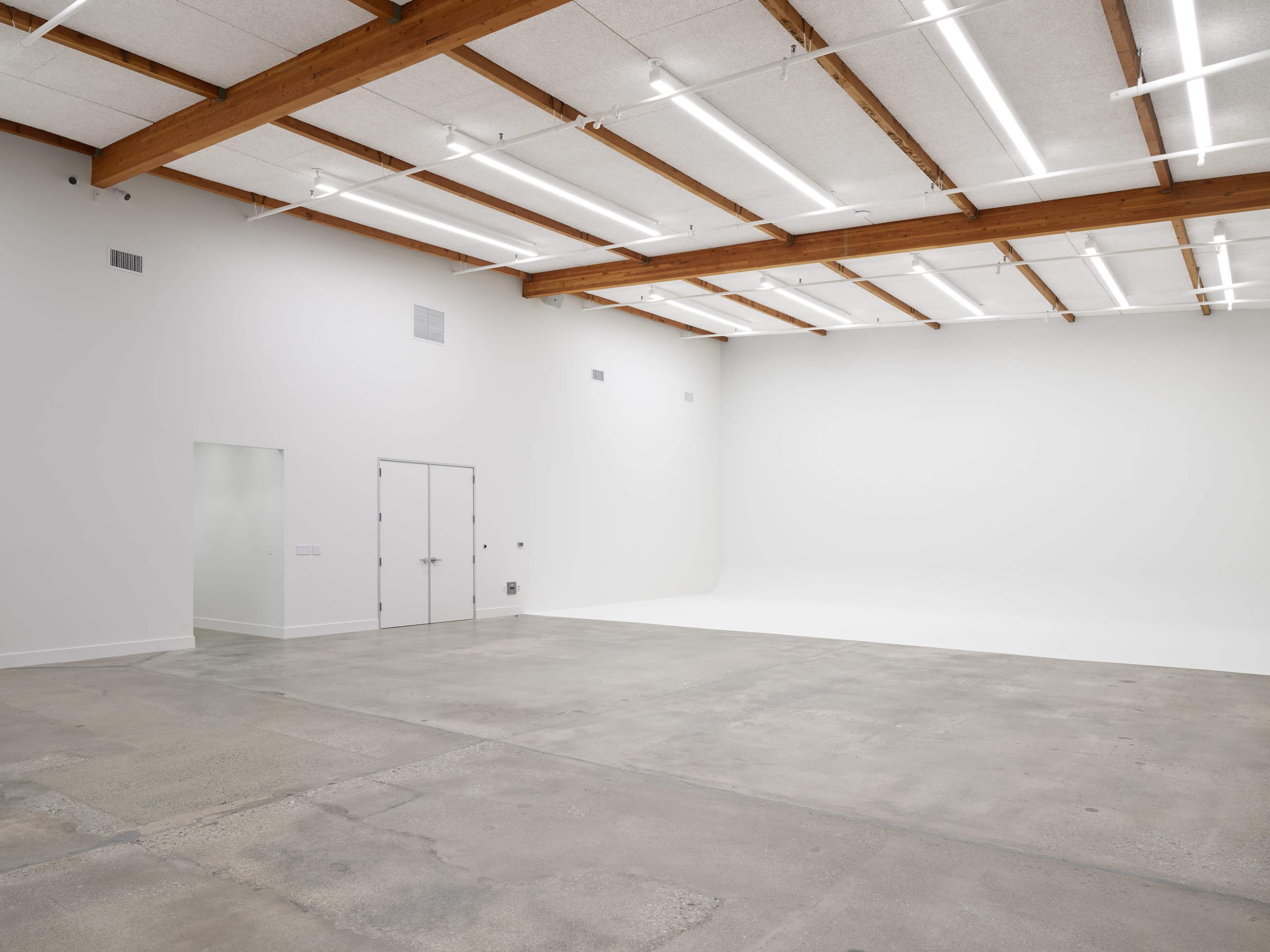 A spacious, empty room with concrete floors and white walls, featuring exposed wooden beams and overhead lighting.