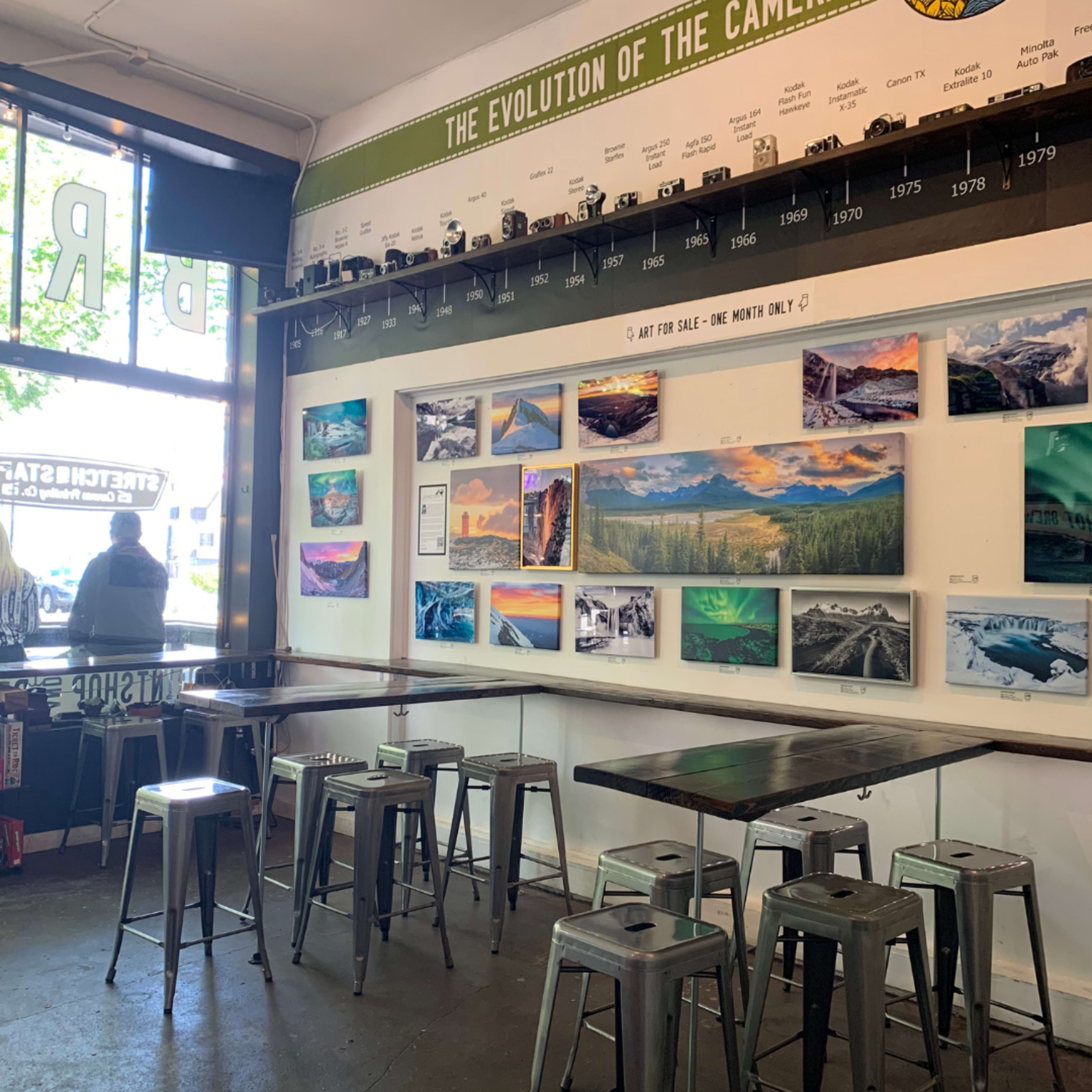 A coffee shop interior featuring metal bar stools, a long table, and a wall displaying photographs alongside a timeline labeled "The Evolution of the Camera."