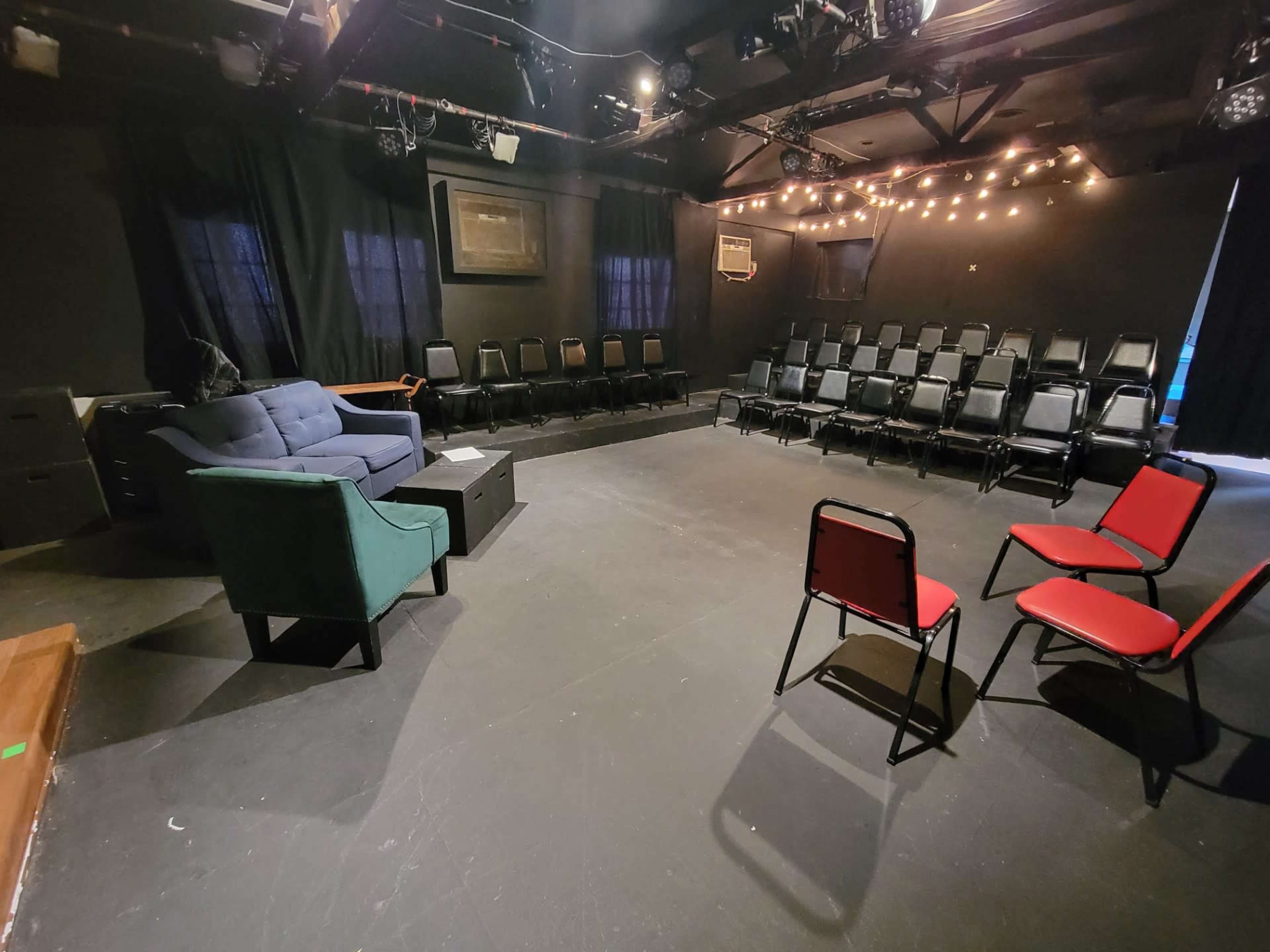 The image shows a small theater setup with a circular arrangement of black chairs, a green and blue couch in the foreground, and dim lighting with string lights overhead.
