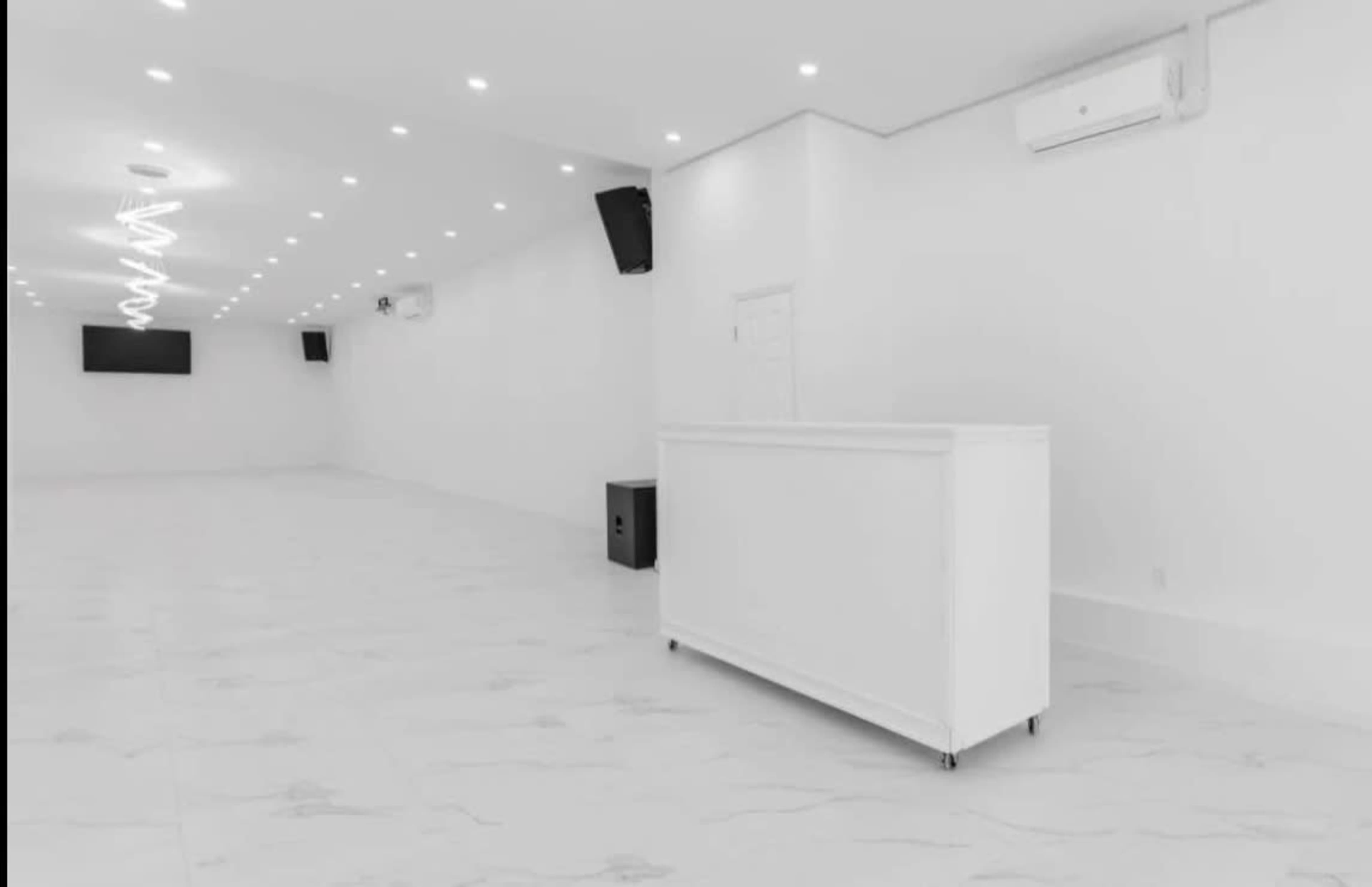 The image shows a spacious, brightly lit room with white walls and floor, featuring a reception area with a white counter and two wall-mounted screens.