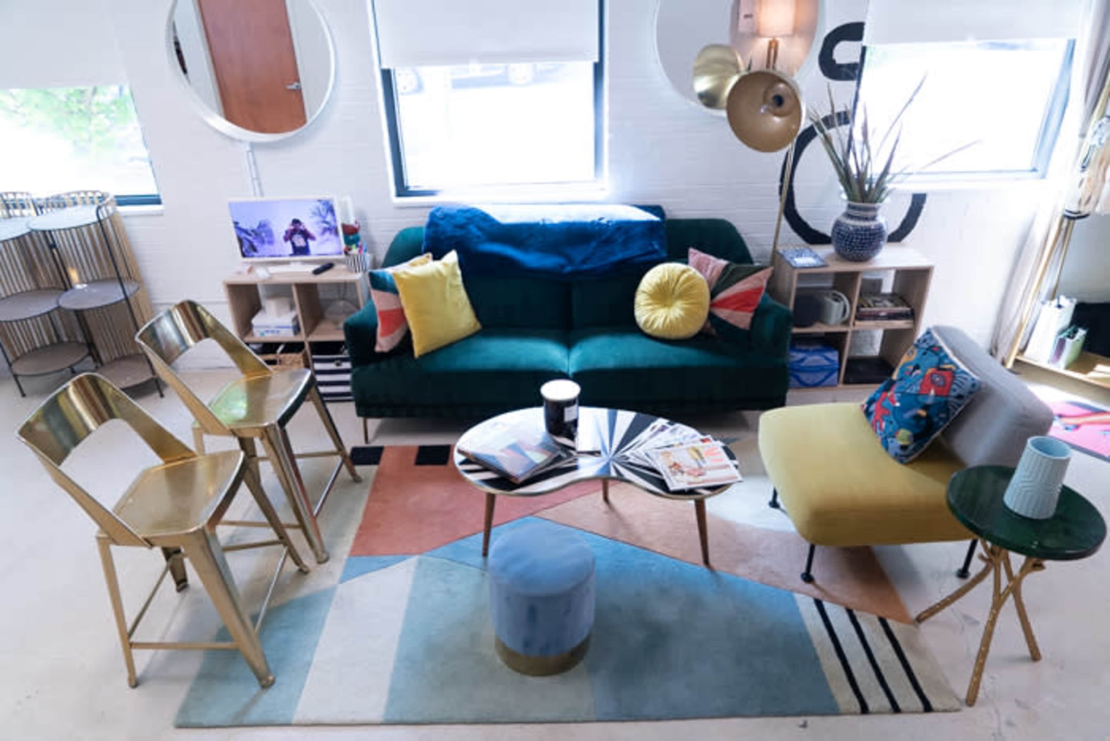 A modern living room with a green sofa, a round coffee table, and a mix of colorful accent chairs, alongside a patterned rug and various decorative items.