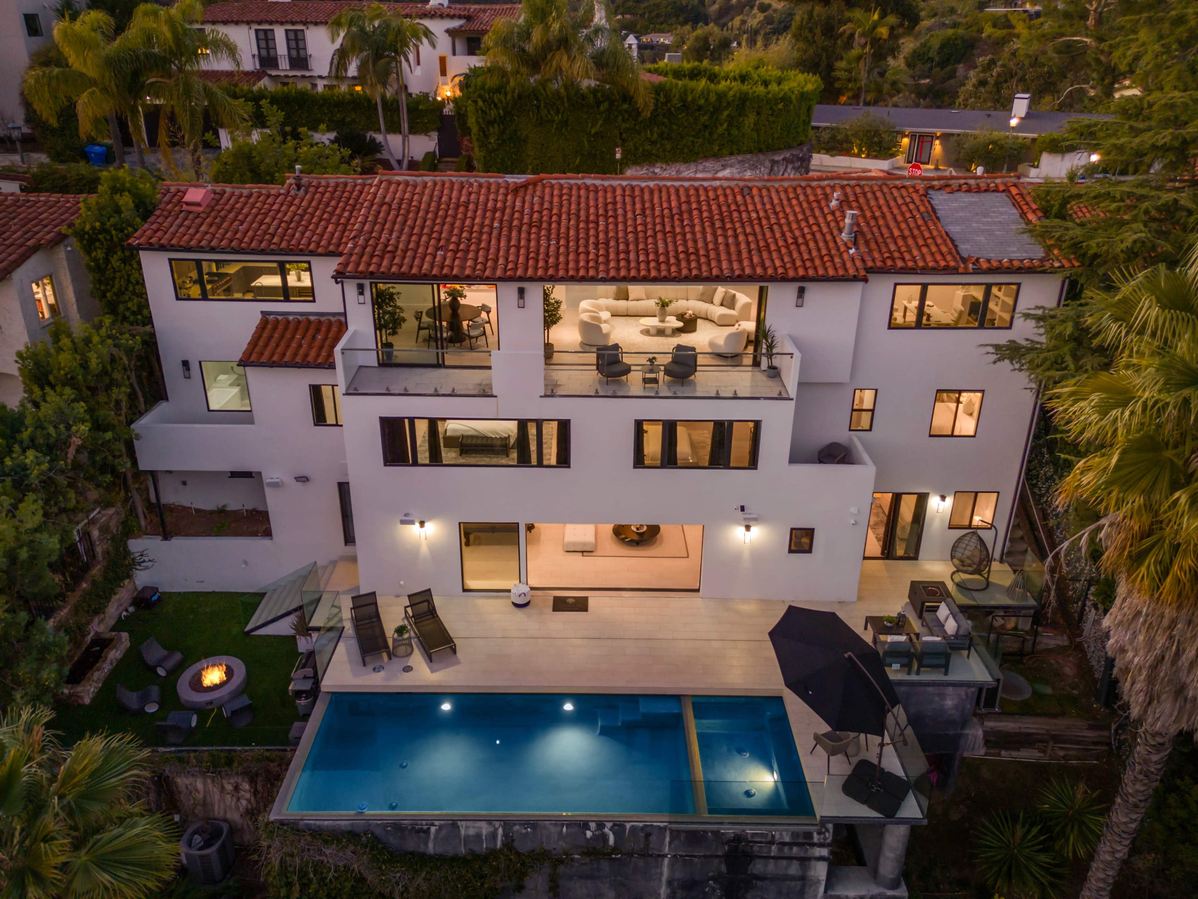 The image shows a modern two-story house with a red-tiled roof, featuring a pool in the backyard and a well-furnished patio area.