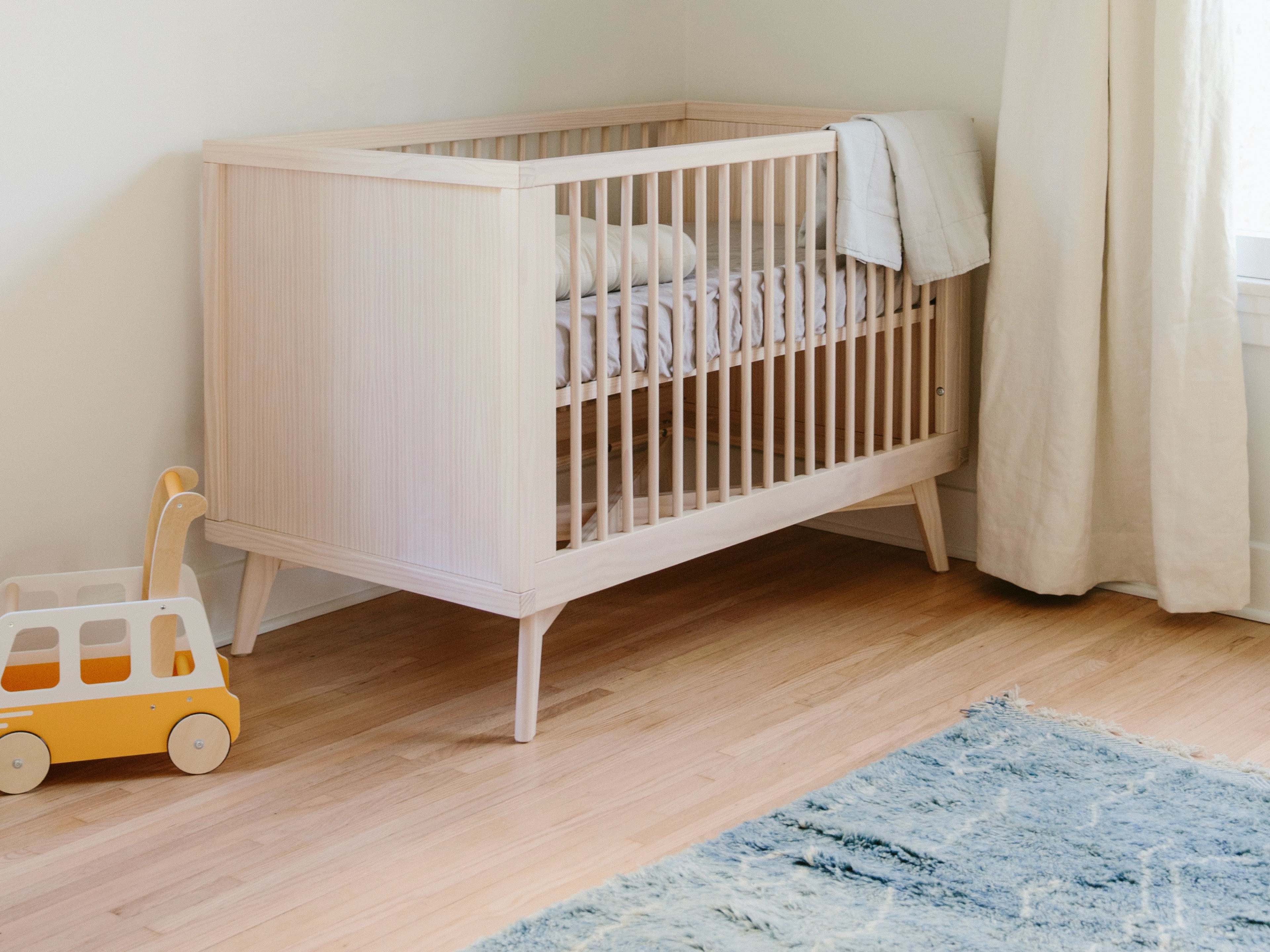 A wooden crib is positioned near a window in a softly lit nursery, accompanied by a yellow toy car on the floor.