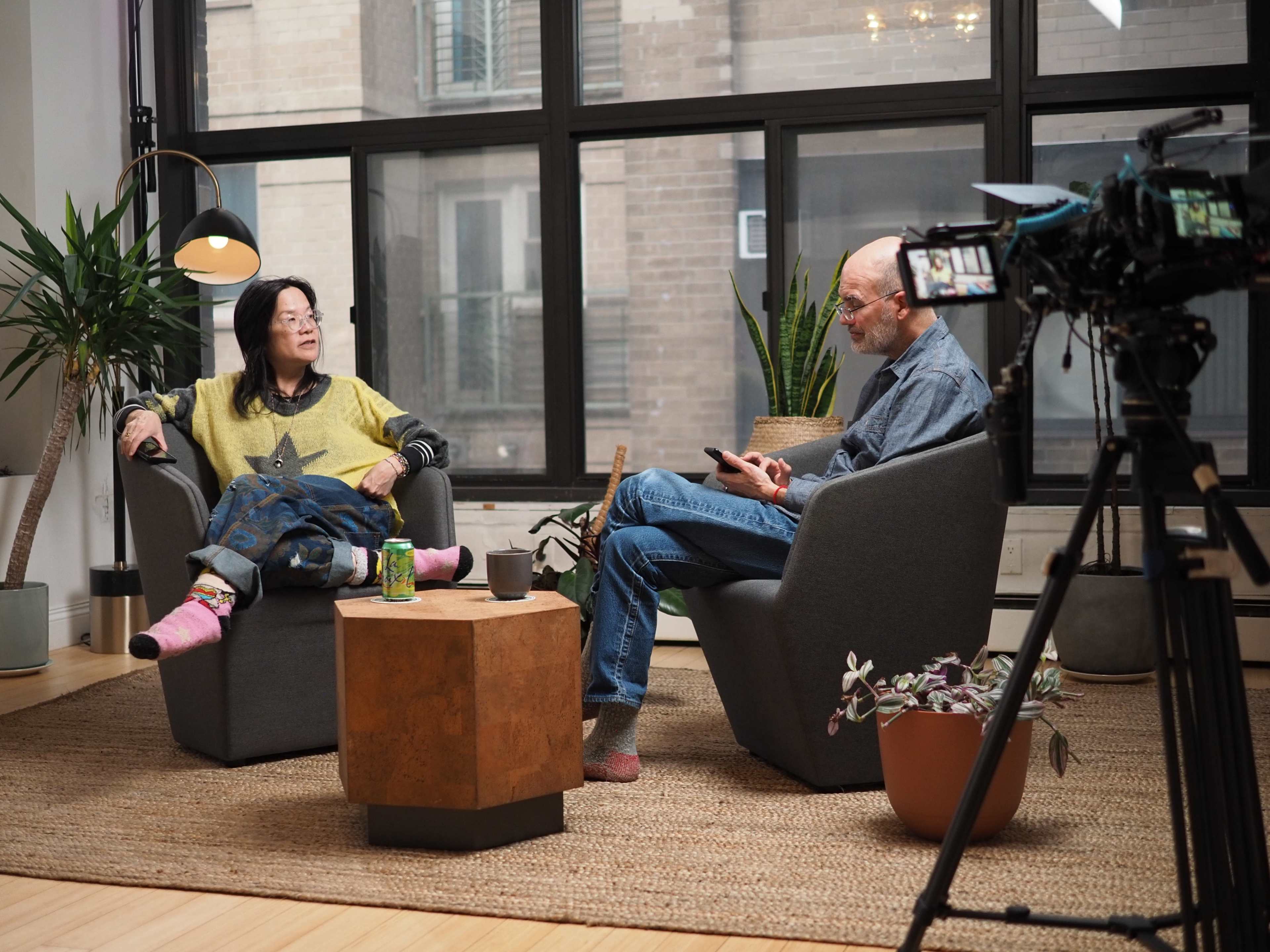A woman and a man sit in modern chairs with a coffee table between them in a well-lit living space featuring plants and large windows.