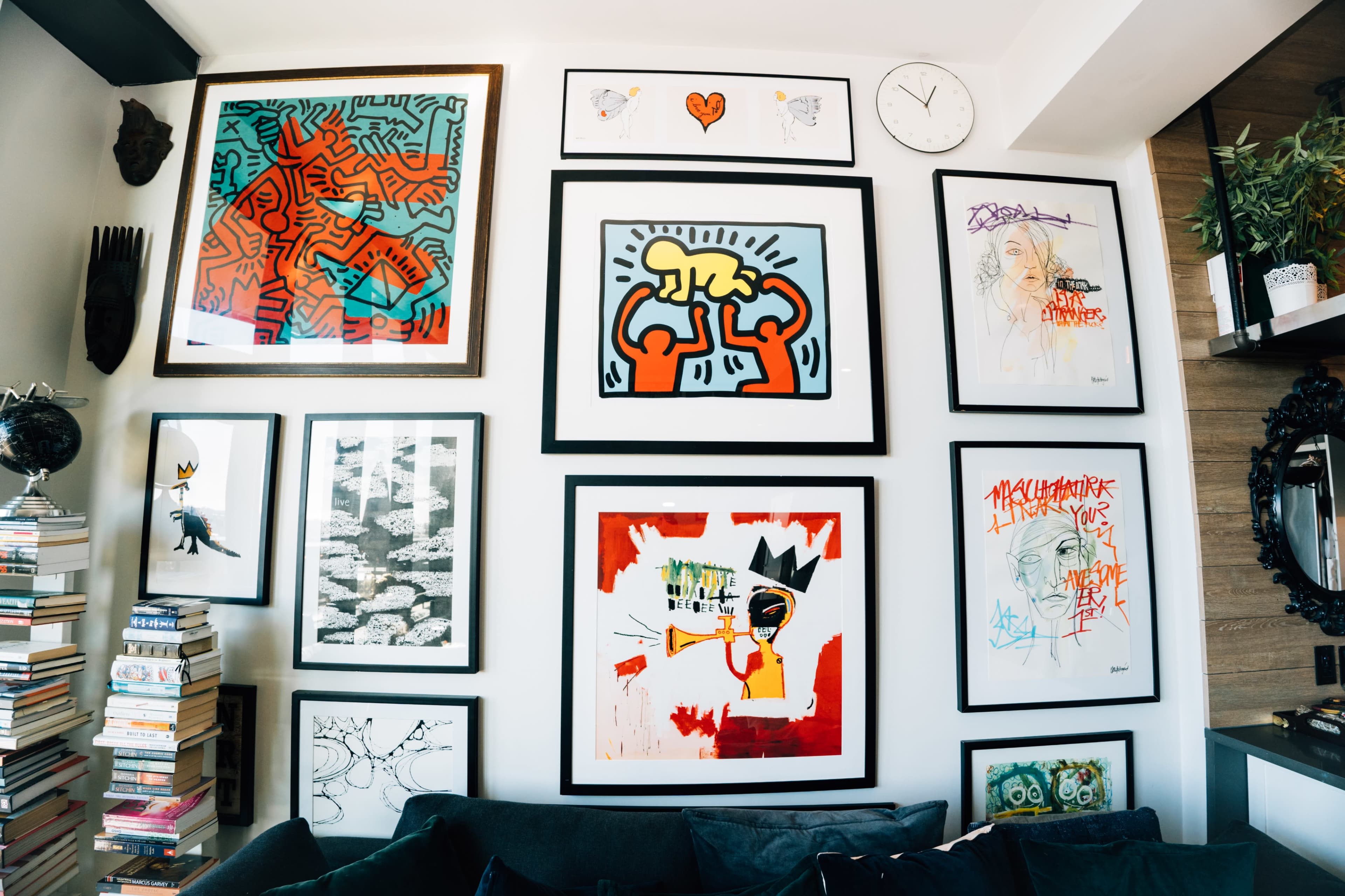 The image showcases a wall adorned with various framed artworks, featuring a mix of abstract and graphic pieces, along with a small clock above.