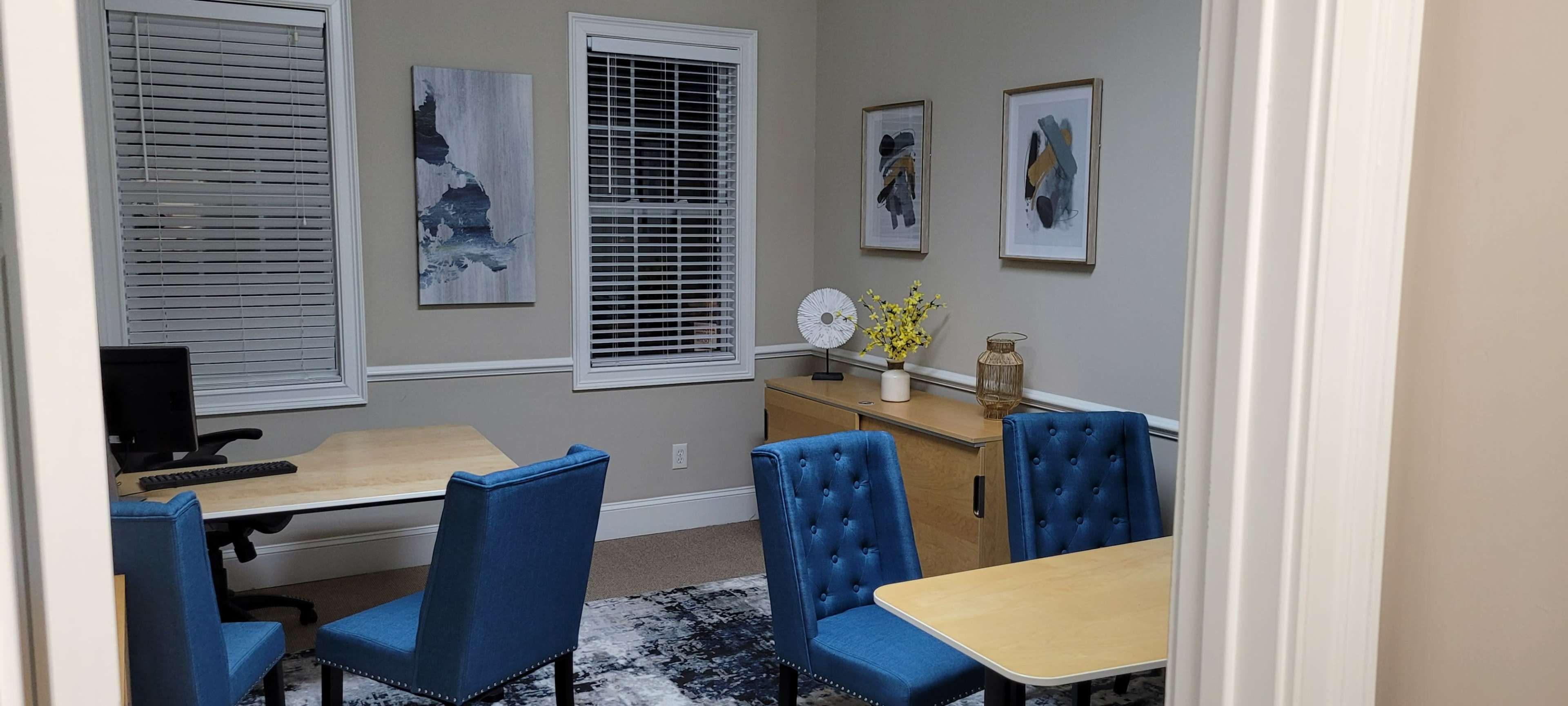 The image shows a well-furnished office space with two blue chairs and wooden tables, featuring artwork on the walls and a window with blinds.