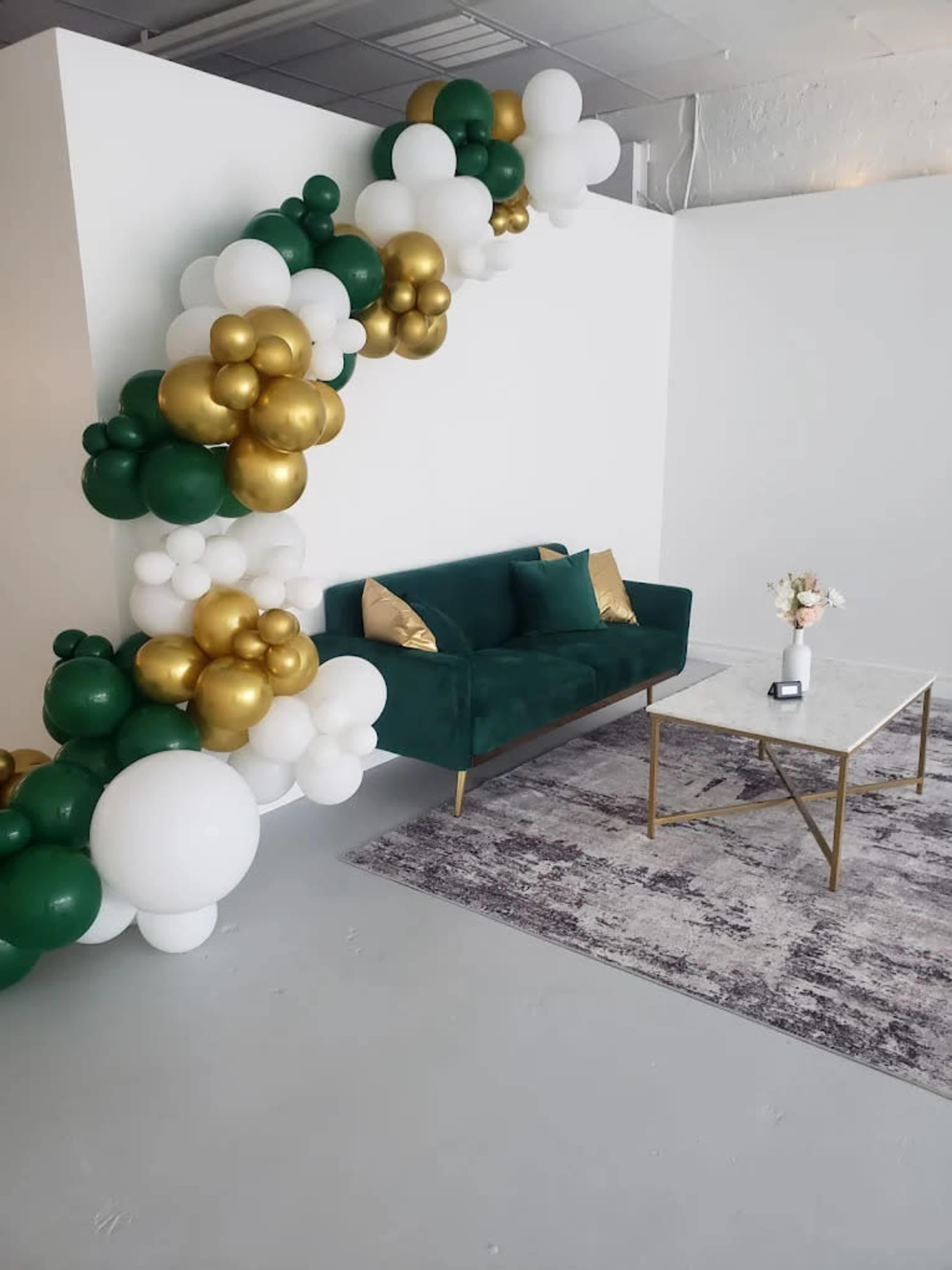 A green sofa with gold and white pillows is positioned next to a marble-top coffee table, under a decorative arrangement of balloons in green, white, and gold along the wall.