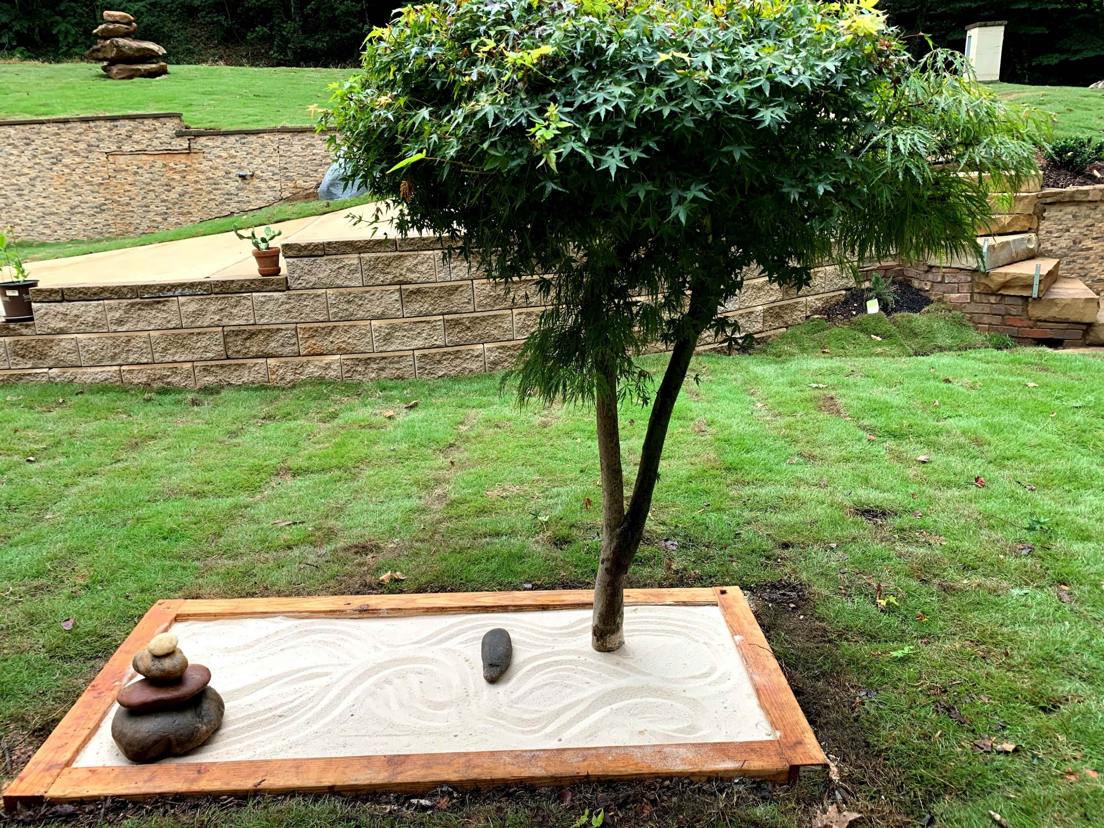 A small tree stands next to a sand garden framed by wooden borders, with stacked stones arranged on one side.