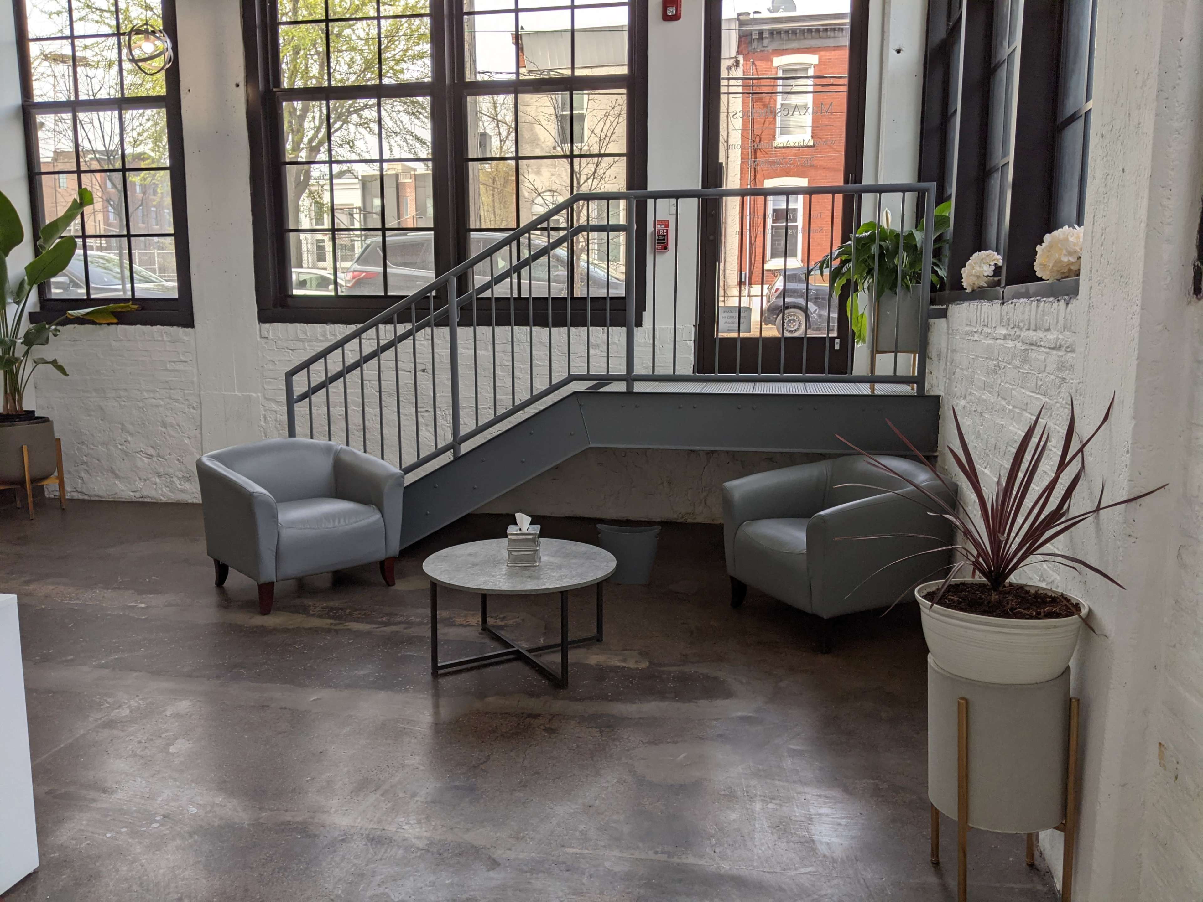The image shows a spacious, well-lit interior with two gray armchairs, a round table, and a staircase leading to an upper level.