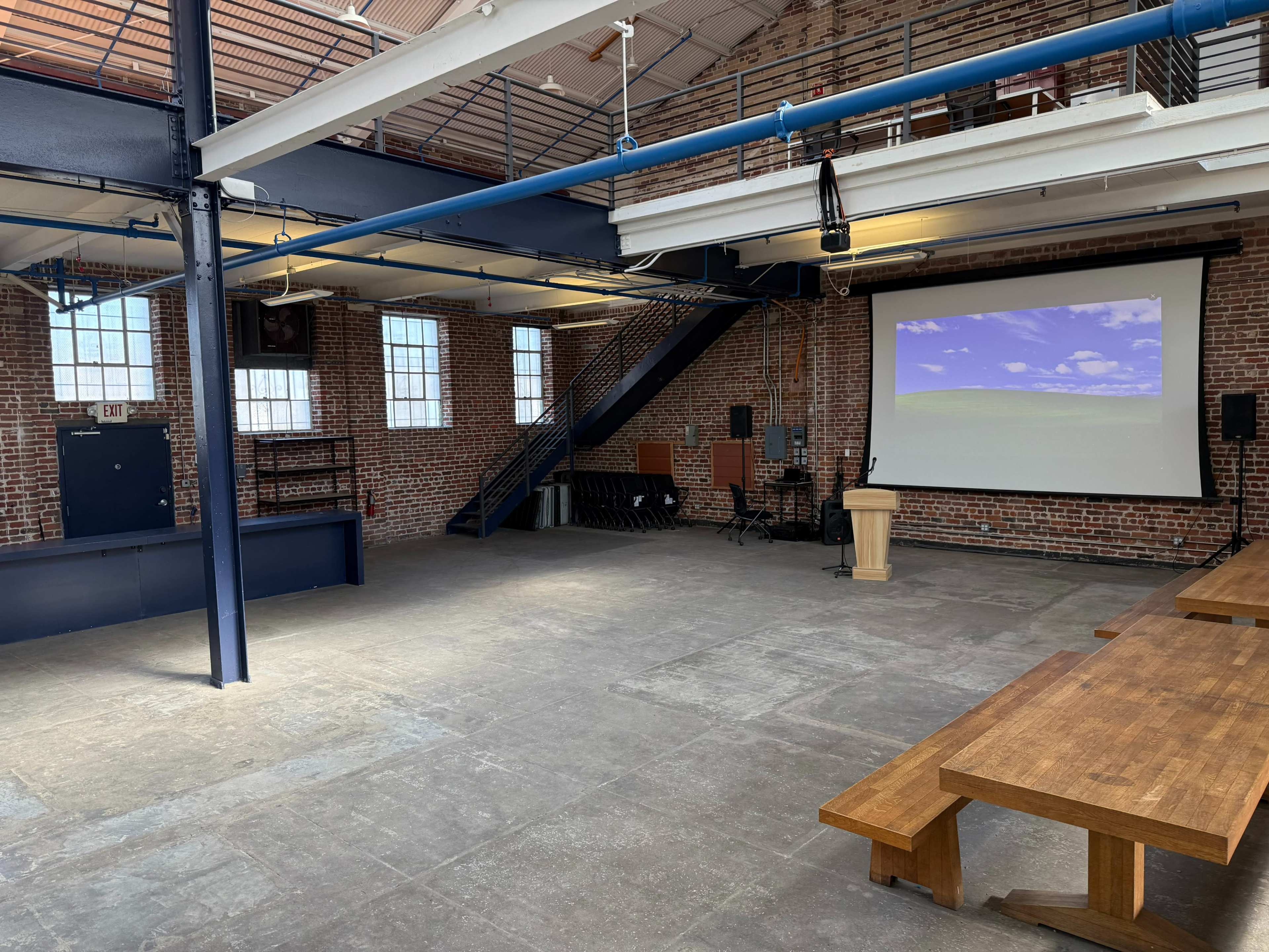 The image shows a spacious, industrial-style room with exposed brick walls, large windows, a stage area, and a wooden podium set up in front of a projection screen.