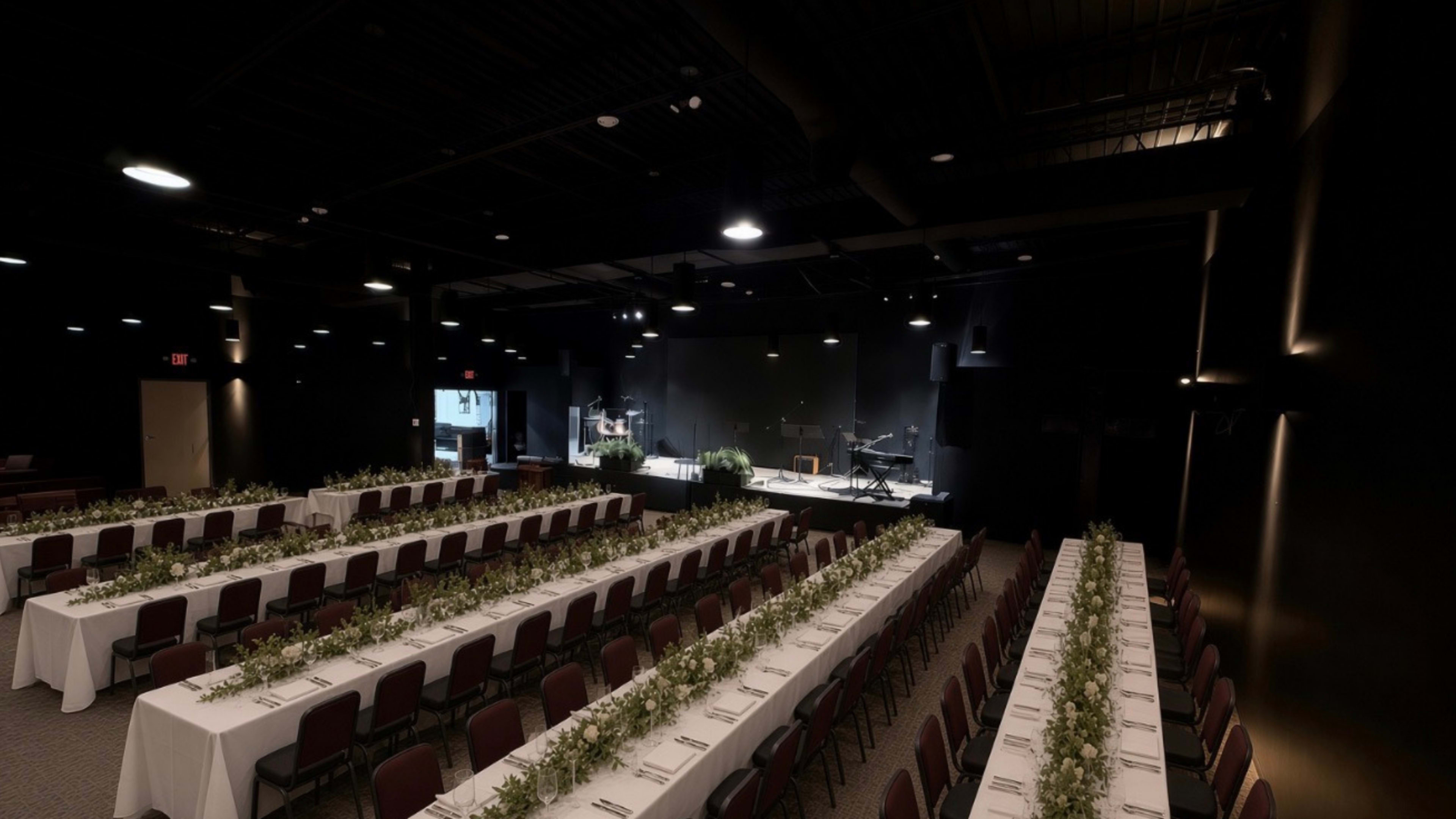 The image shows a spacious indoor venue set up for an event, featuring multiple long tables with white tablecloths arranged in rows and greenery decorations along the center, facing a small stage with musical instruments.