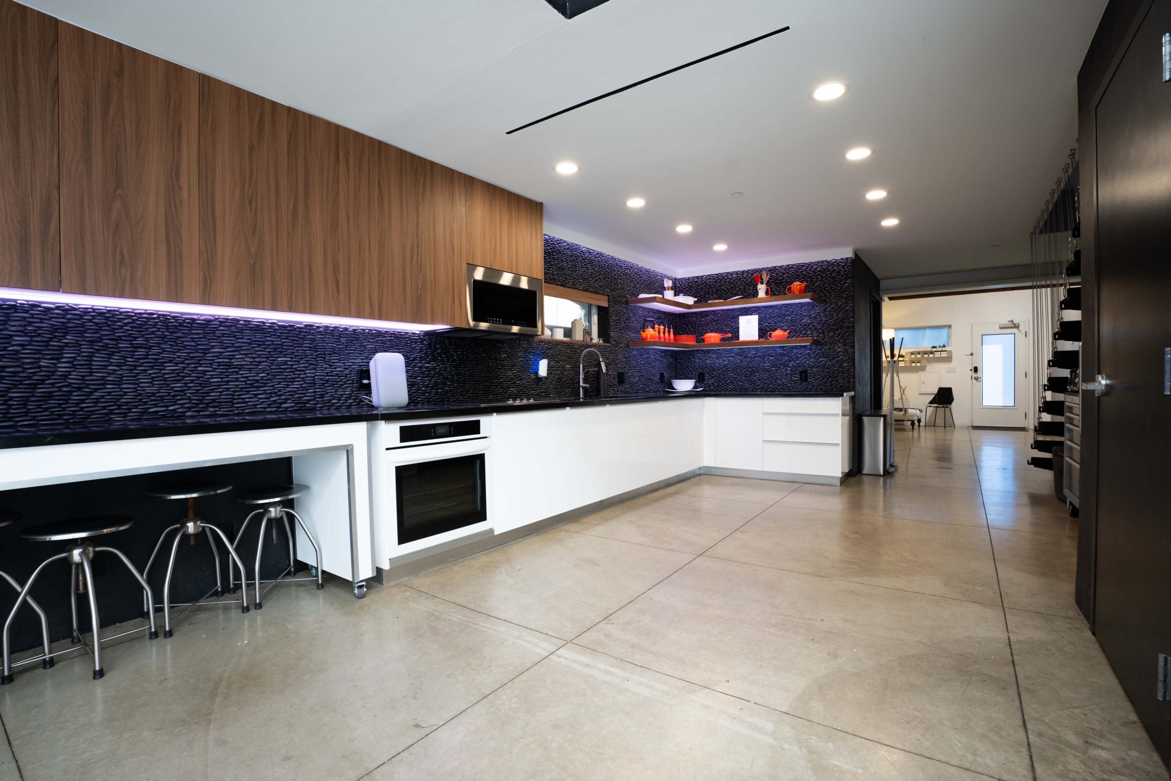 The image shows a modern kitchen with sleek white cabinetry, dark countertops, and wooden accents, featuring a minimalist design and open layout.