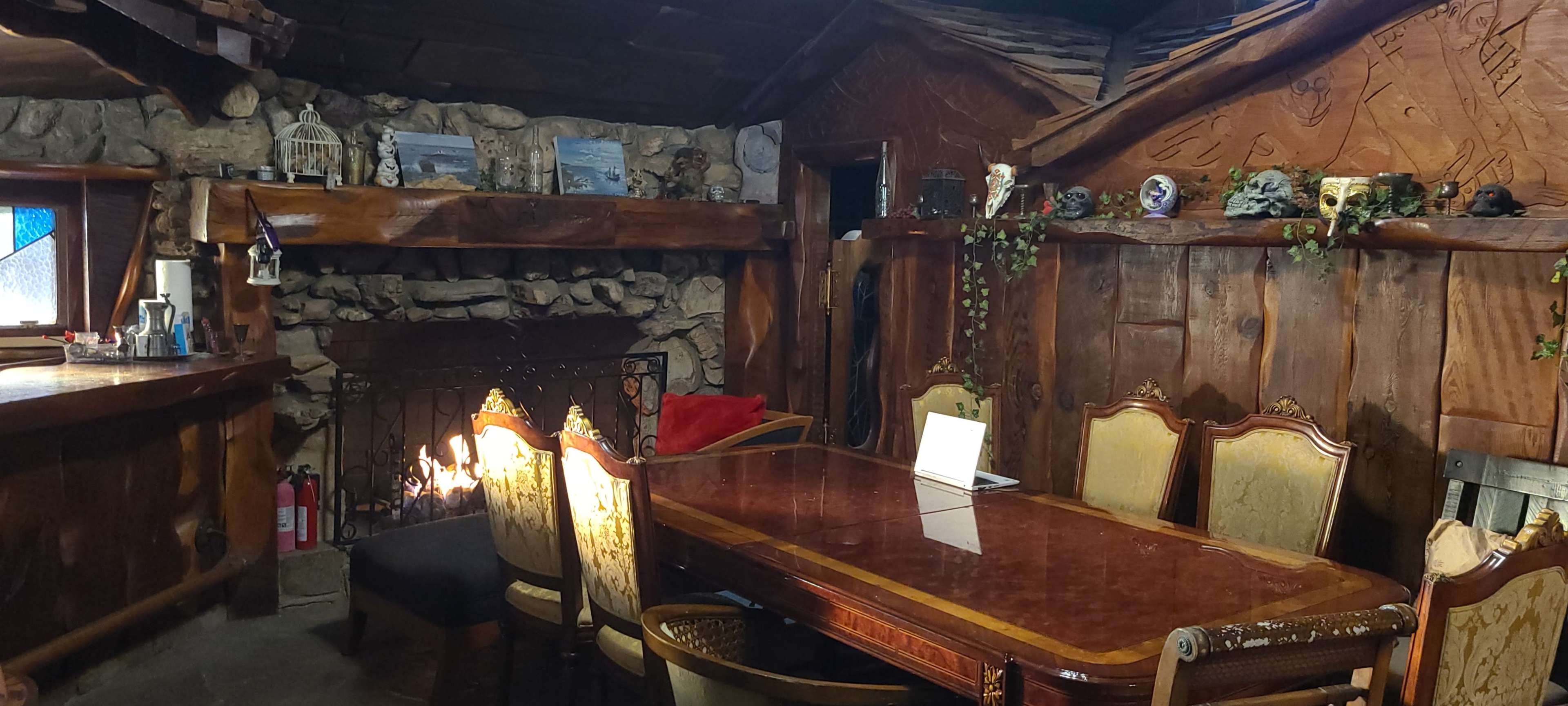A rustic dining area features a wooden table surrounded by upholstered chairs, with a stone fireplace and carved wood walls.
