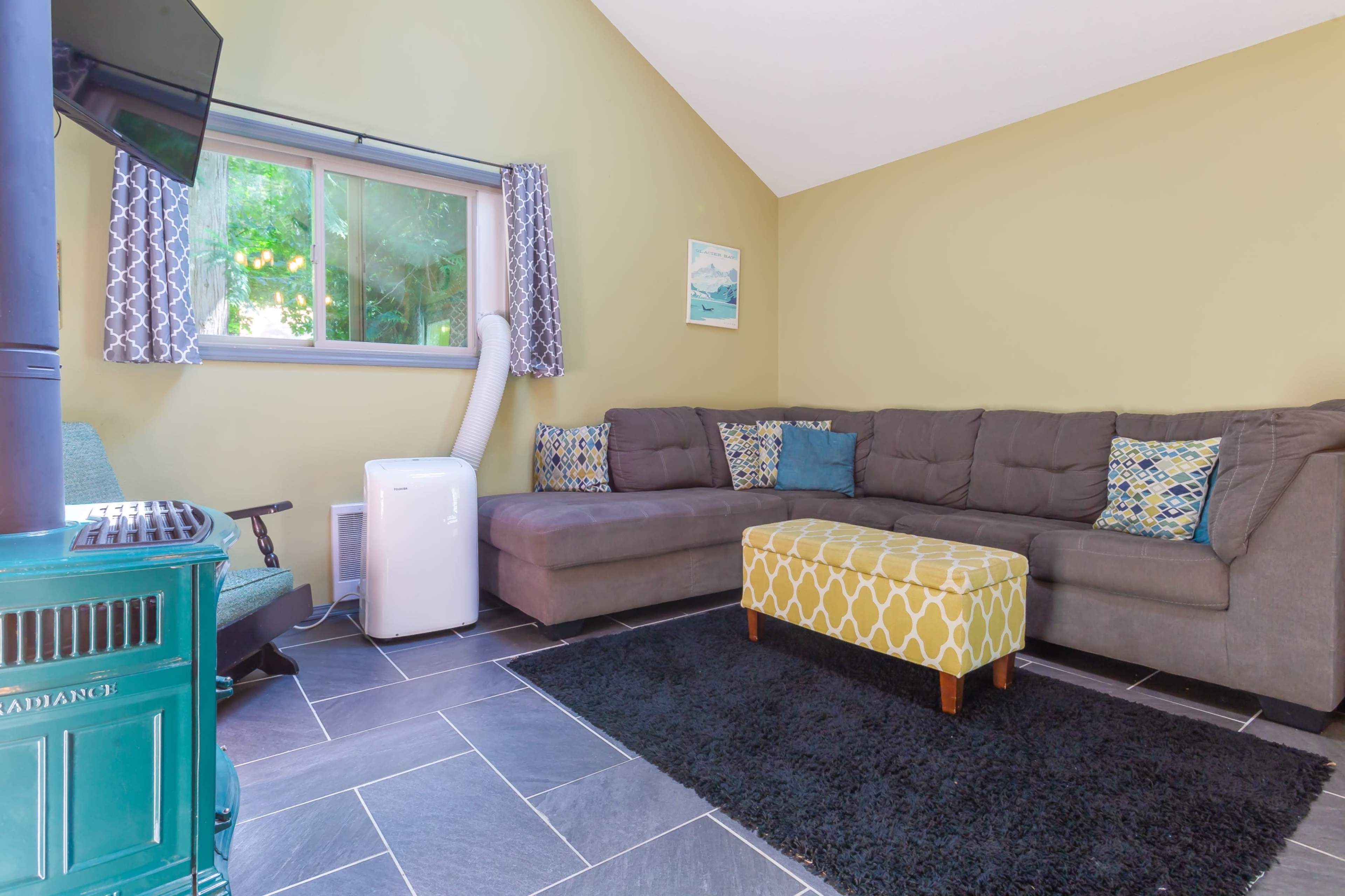 A cozy living room with a sectional sofa, a yellow ottoman, a black rug, and a window allowing natural light to come in.