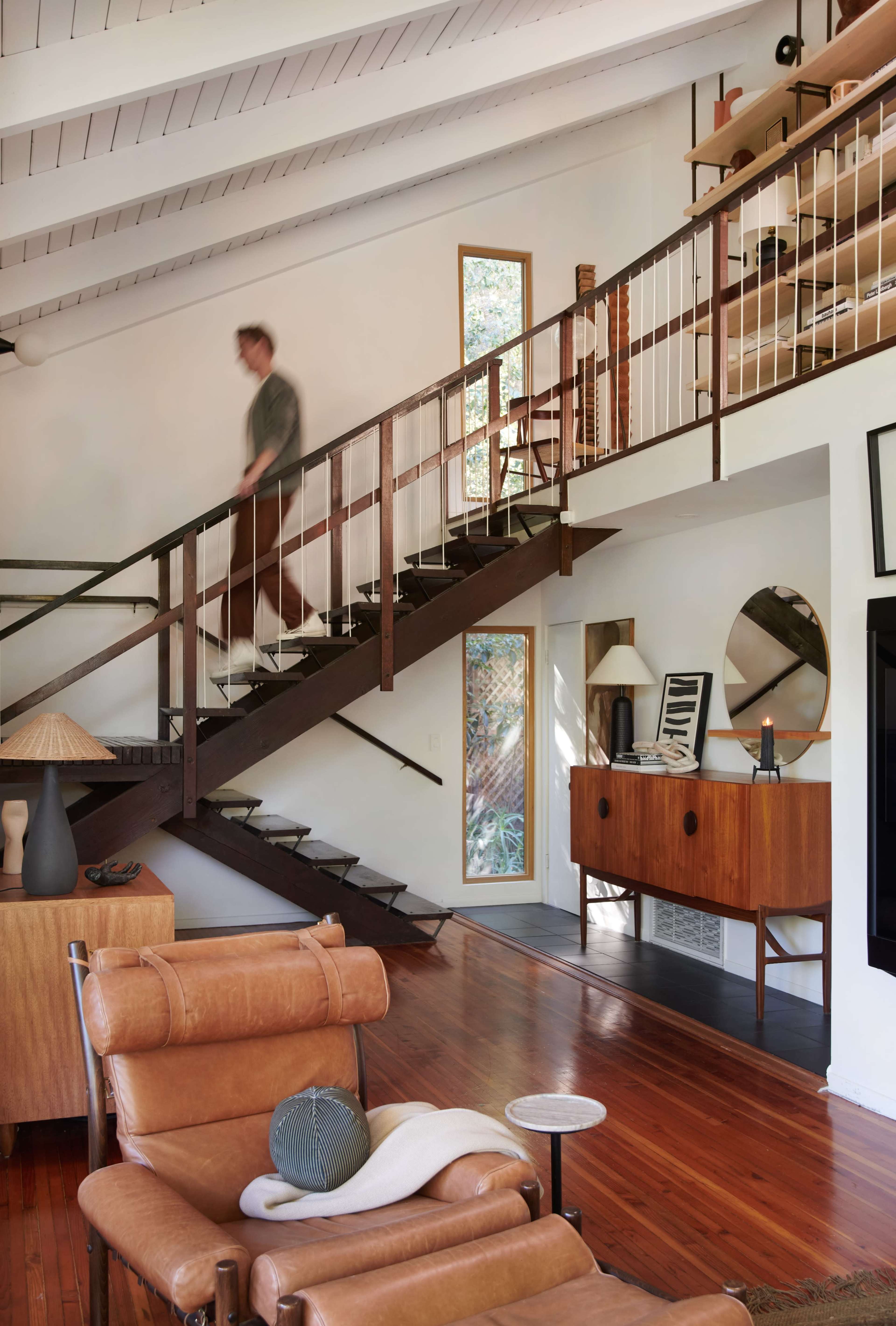 A person walks down a modern staircase in a spacious living room featuring mid-century furniture and large windows.