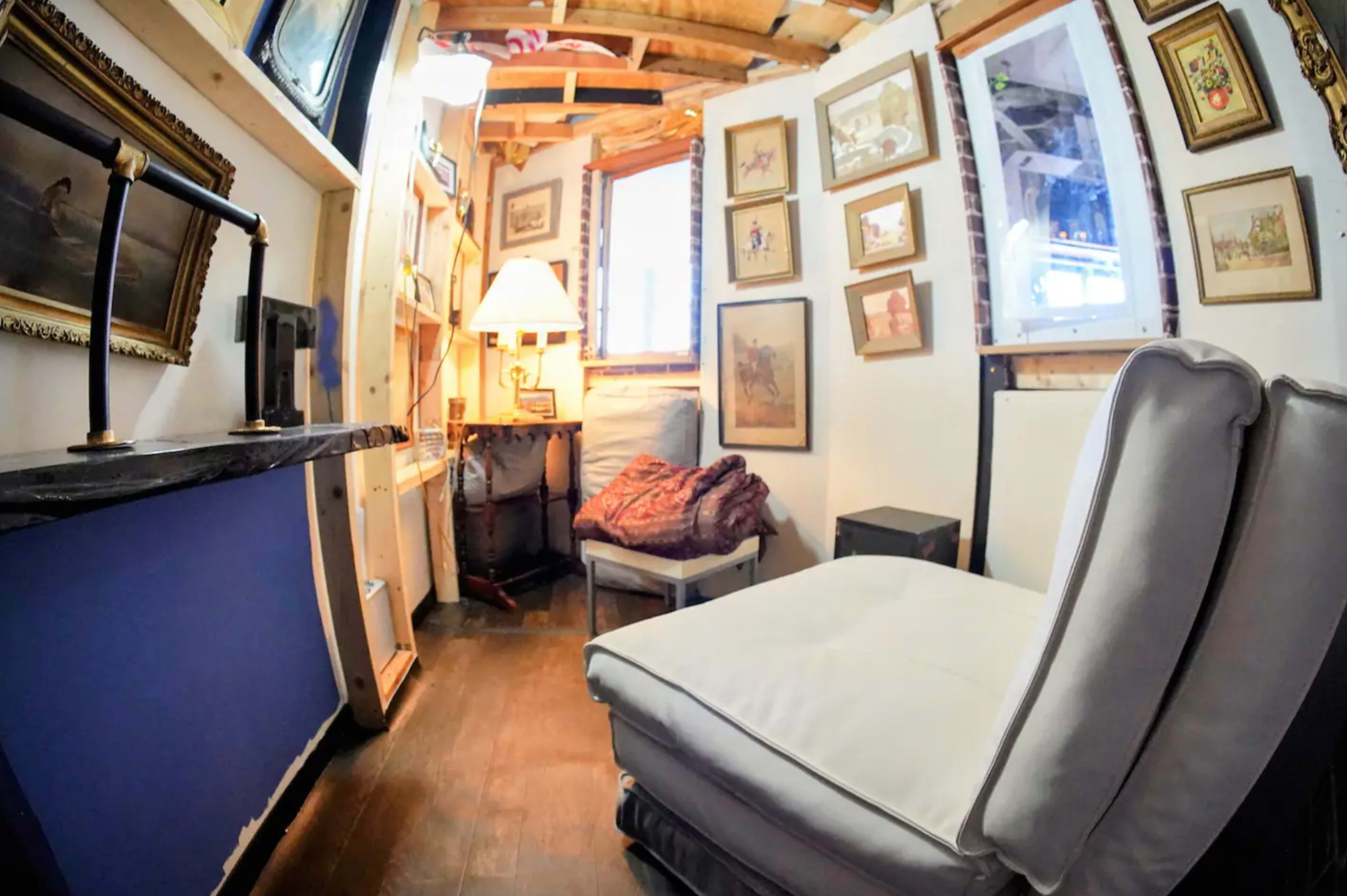 A small, cozy room with a white chair, framed artwork on the walls, and a lamp in a corner near a window.