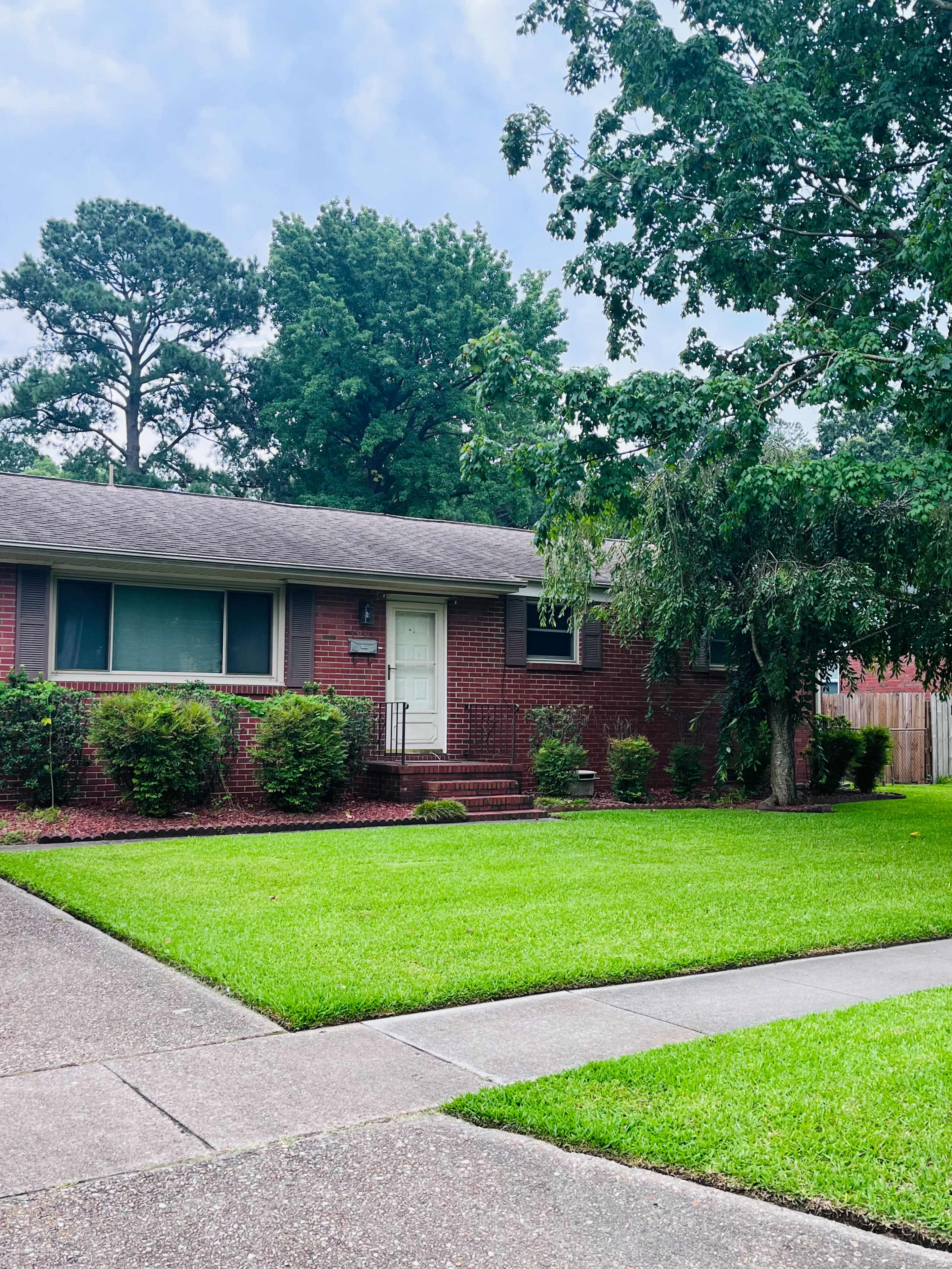 A red brick house with a white door is situated on a well-maintained lawn, surrounded by shrubs and trees.