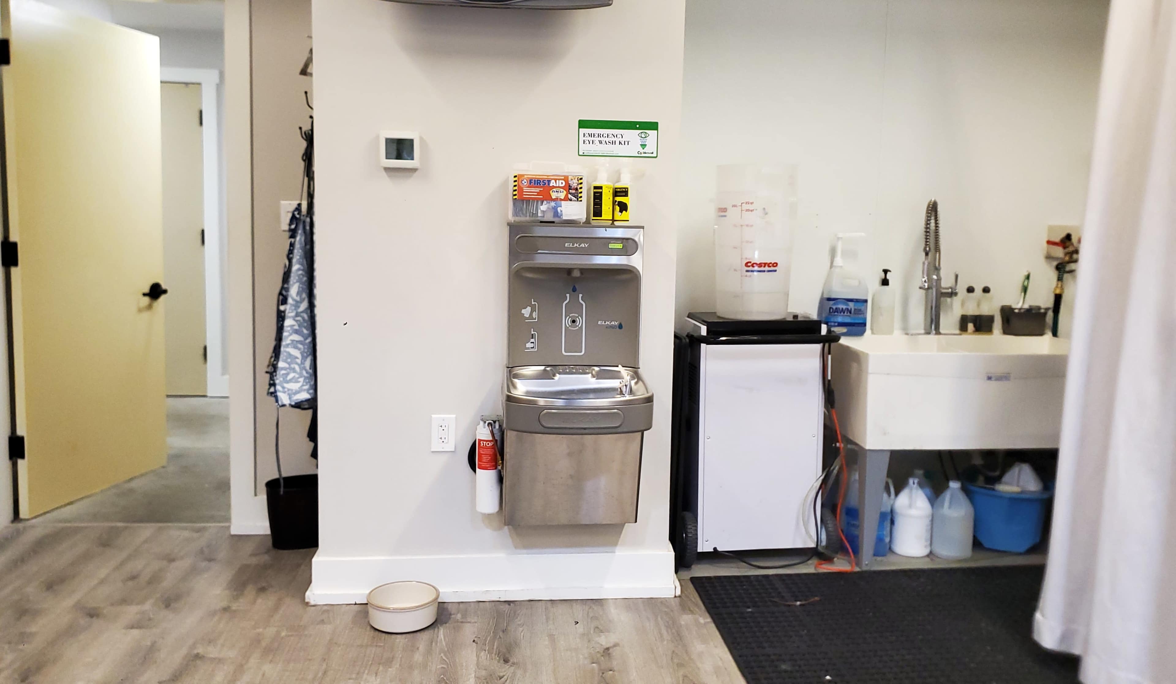 The image shows a water fountain mounted on a wall beside a sink and a black mat, with a door leading to another room in the background.